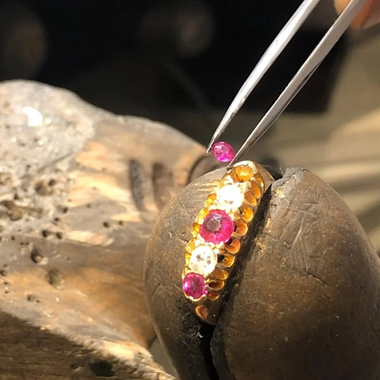 A jewelry craftsman setting pink and white gemstones into a gold ring with tweezers, on a  wooden workbench.