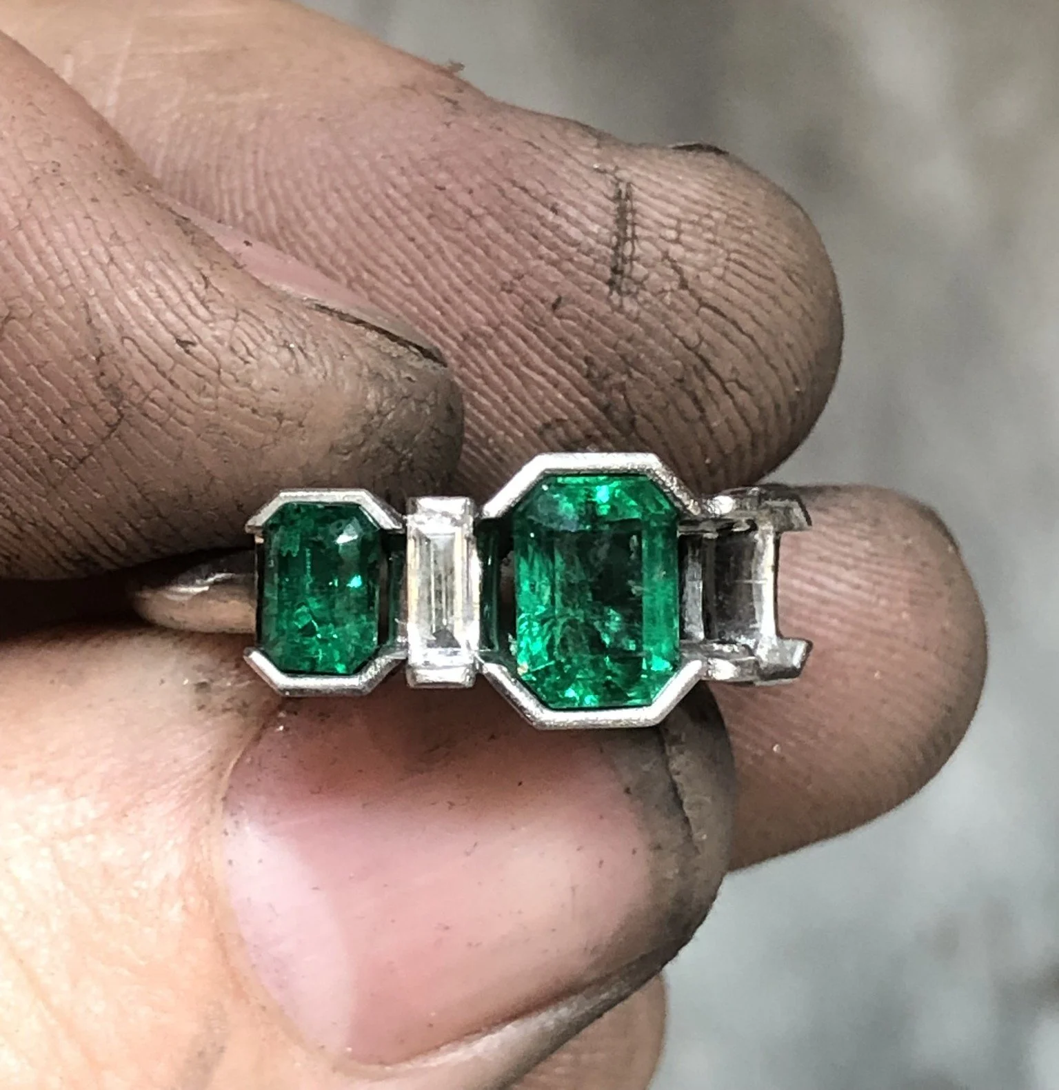 Close-up of a white gold ring with two large emerald-cut green gemstones , missing a stone.