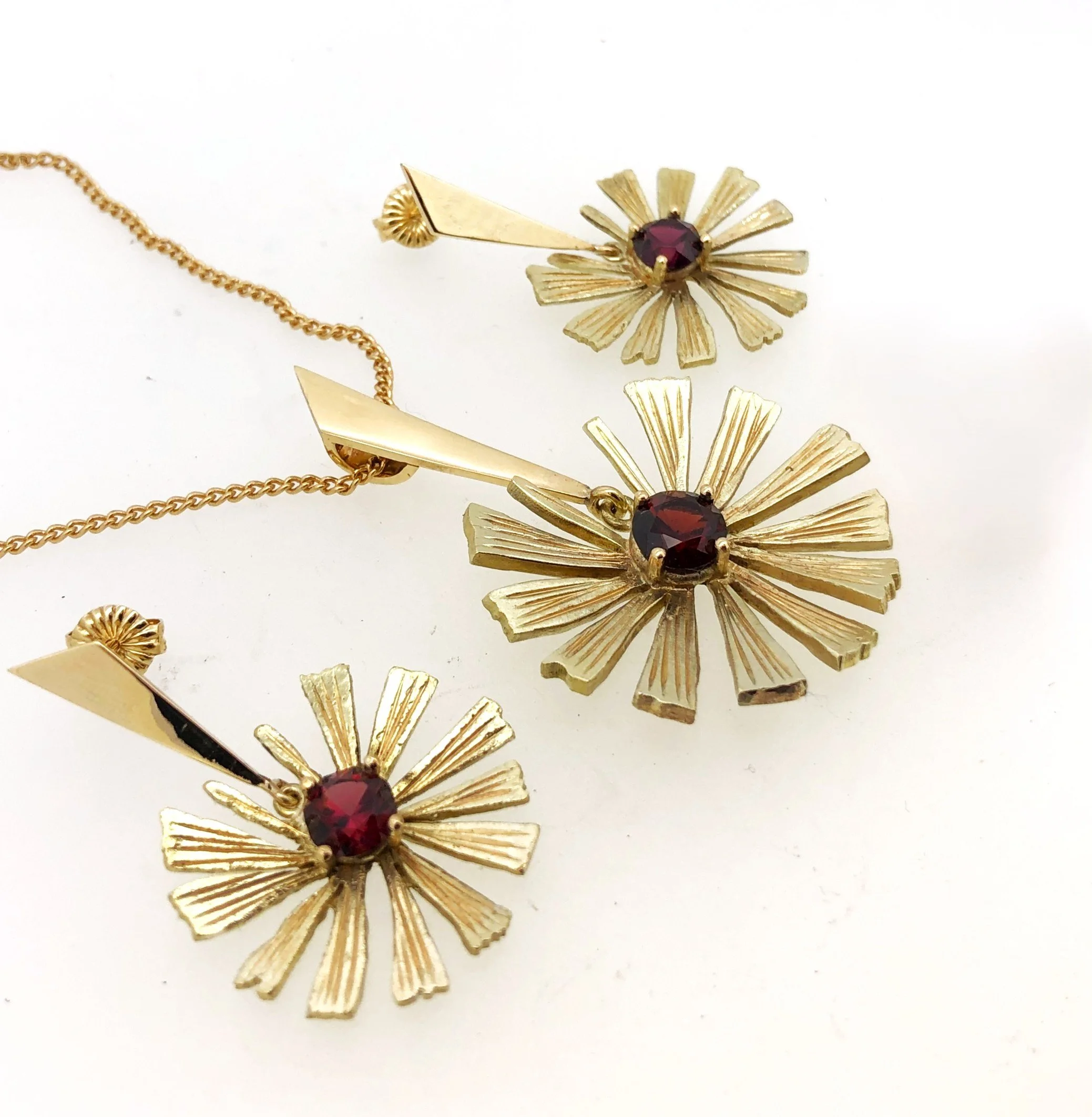 Gold jewelry pieces including a pendant with a red gemstone and matching earrings with a sunburst design, displayed on a white background.