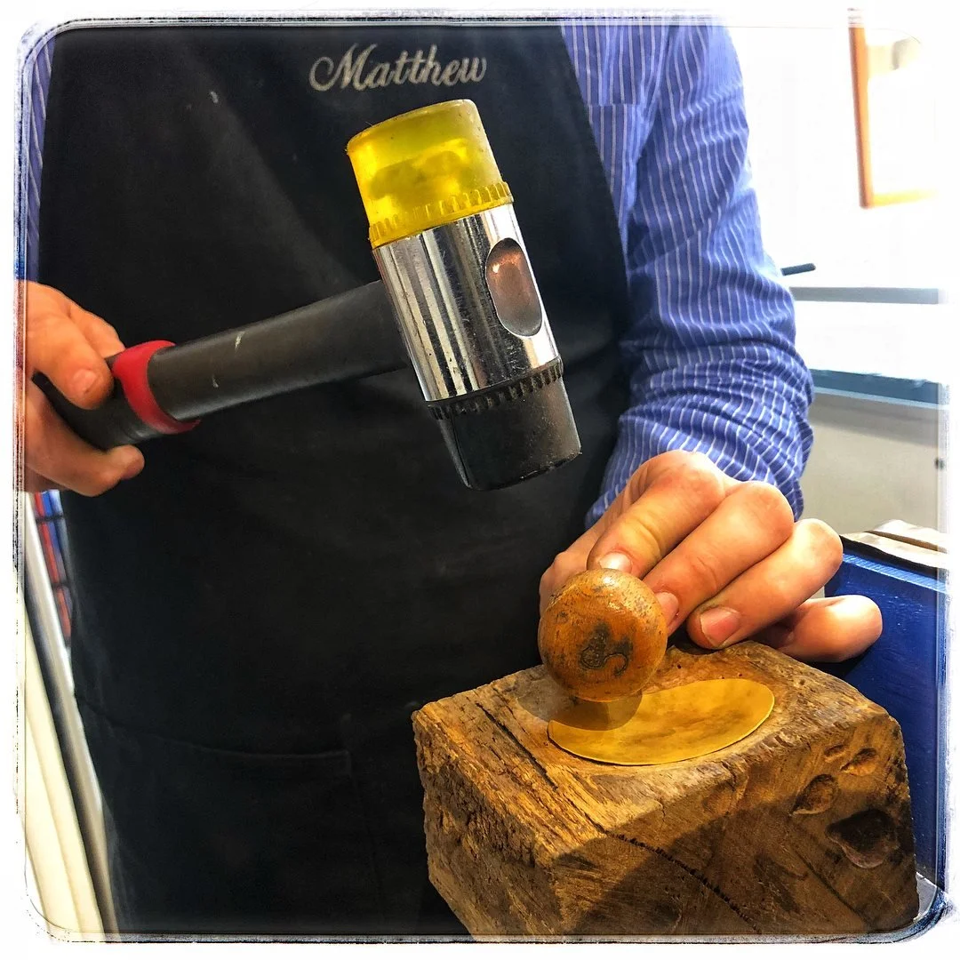 Jeweller using a hammer and a mallet on a piece of wood to shape a rounded metal object.