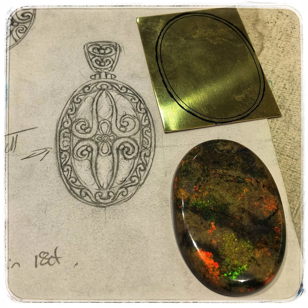 Sketch of an ornate oval-shaped pendant necklace with intricate swirling patterns, a gemstone to go into the pendant and a flat piece of rolled gold, on paper with handwritten notes.