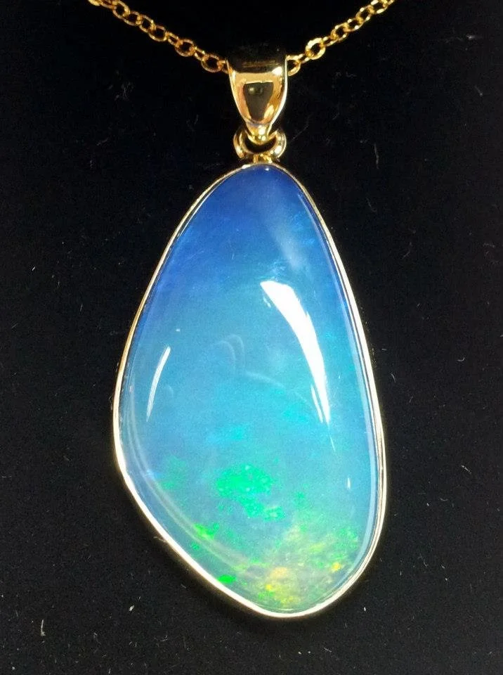 Gold chain necklace with a large teardrop-shaped opal pendant, displaying blue and green hues, hanging against a black background.