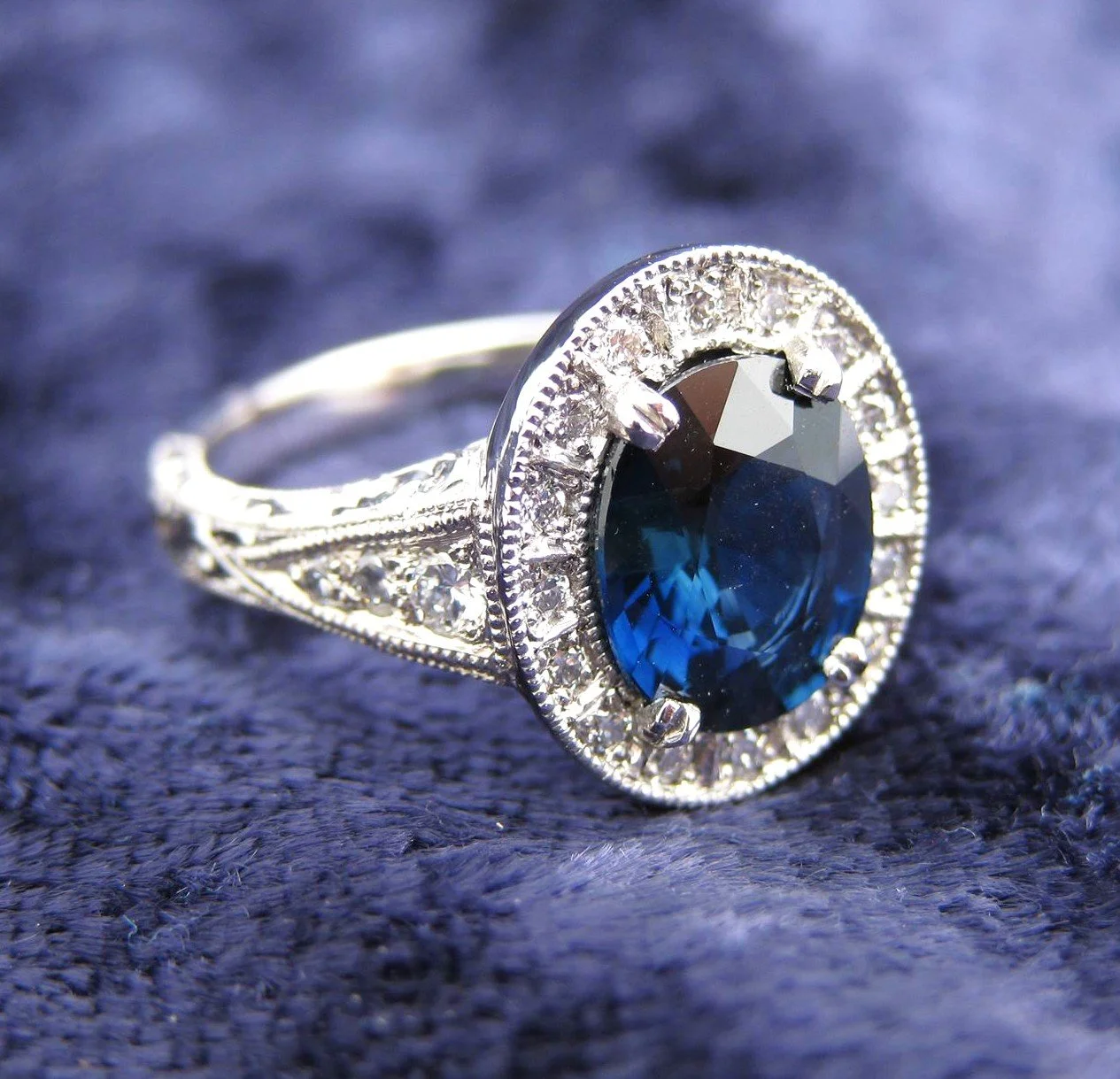 A silver ring with a large deep blue gemstone surrounded by small clear diamonds, set on a textured blue fabric background.