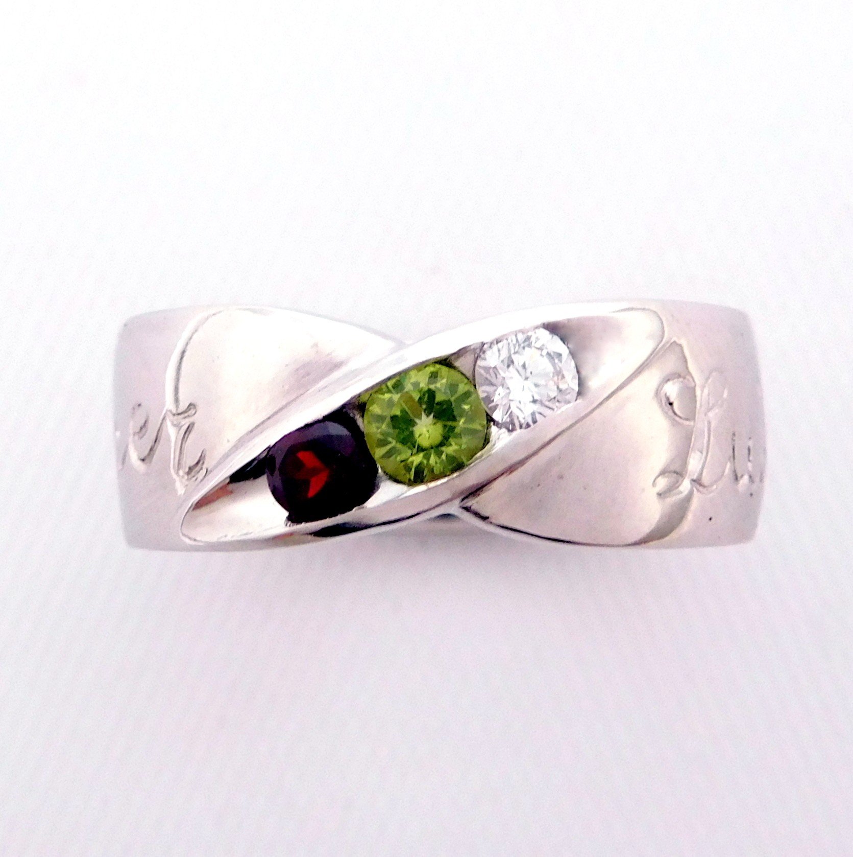 A white background with a metallic ring featuring four gemstones: a green peridot, a clear diamond, a dark purple amethyst, and a red garnet.