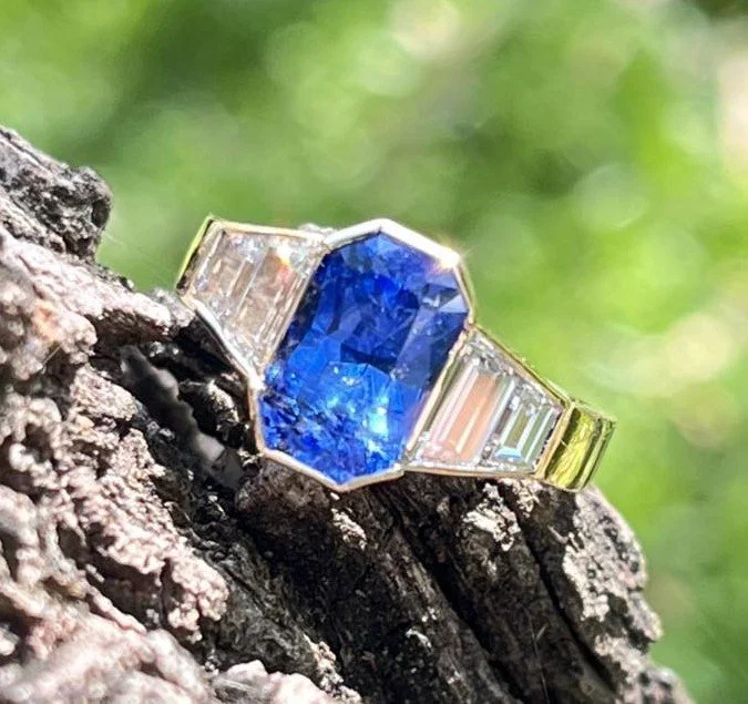 A gold ring with a large, rectangular blue gemstone and smaller baguette-cut diamonds on each side, resting on tree bark with a blurred green background.
