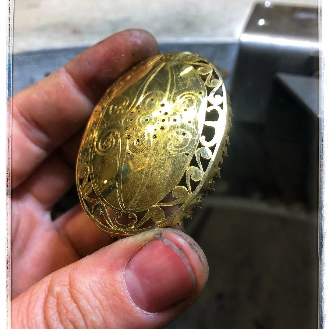 Close-up of a person's hand holding a shiny, gold-colored, oval-shaped decorative item with intricate engraved patterns and filigree edges.