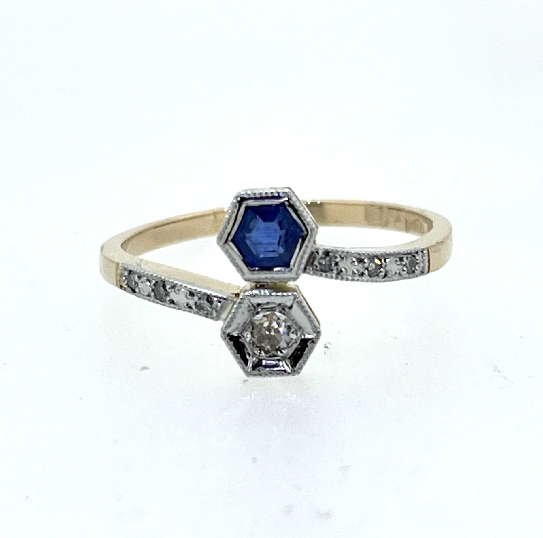 Close-up of a gold ring with a large, blue, hexagonal gemstone and smaller diamonds around it, atop a white surface.