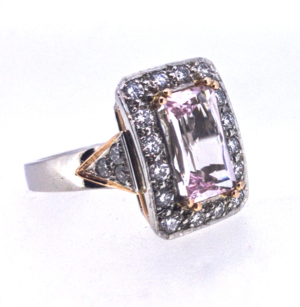 A jewelry ring with a large rectangular pink gemstone in the center, surrounded by smaller clear stones, and a band with gold accents on the sides.