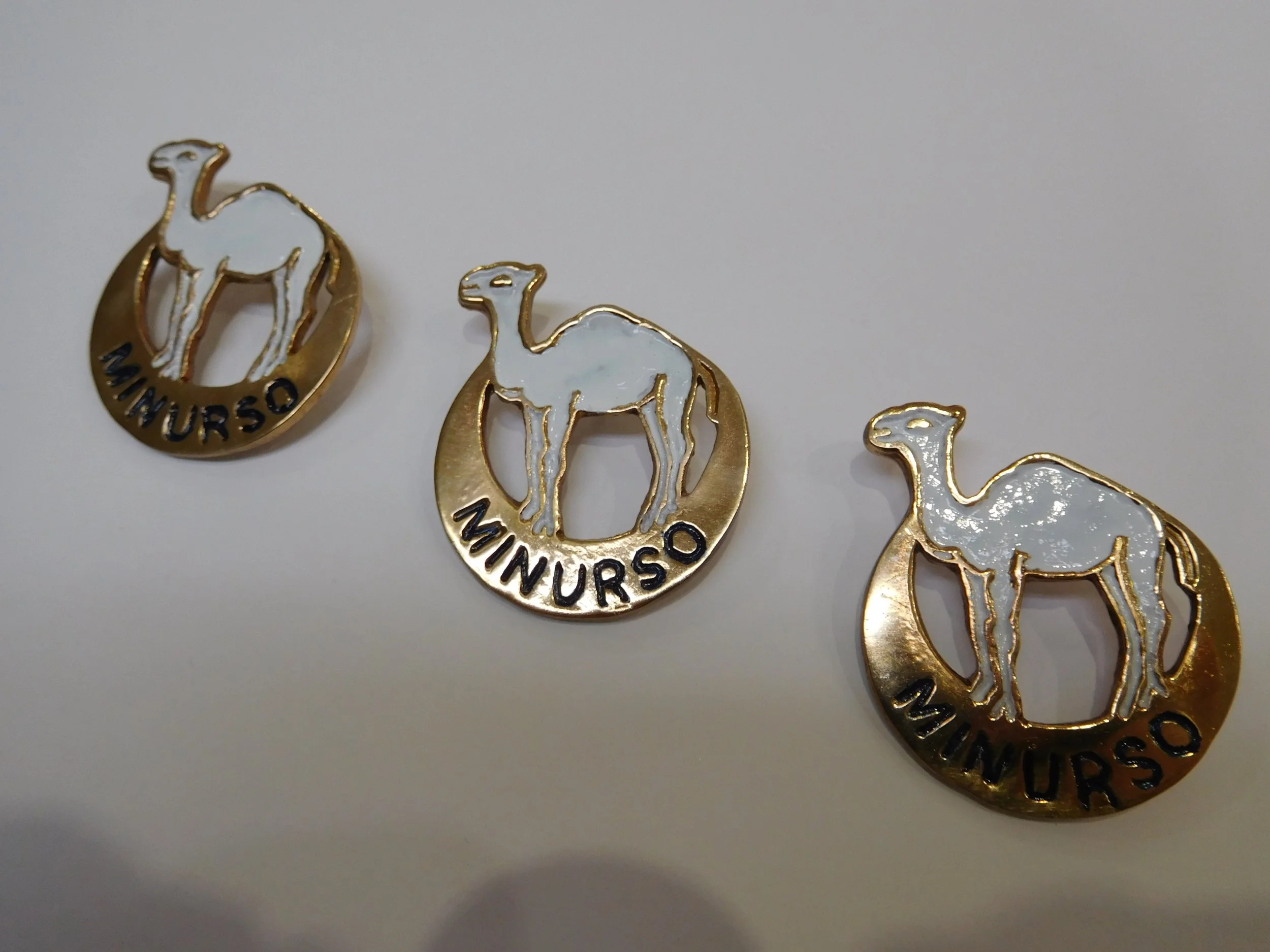 Group of gold and white camel-shaped pins with the word 'MUNIRSO' on the bottom, military items.