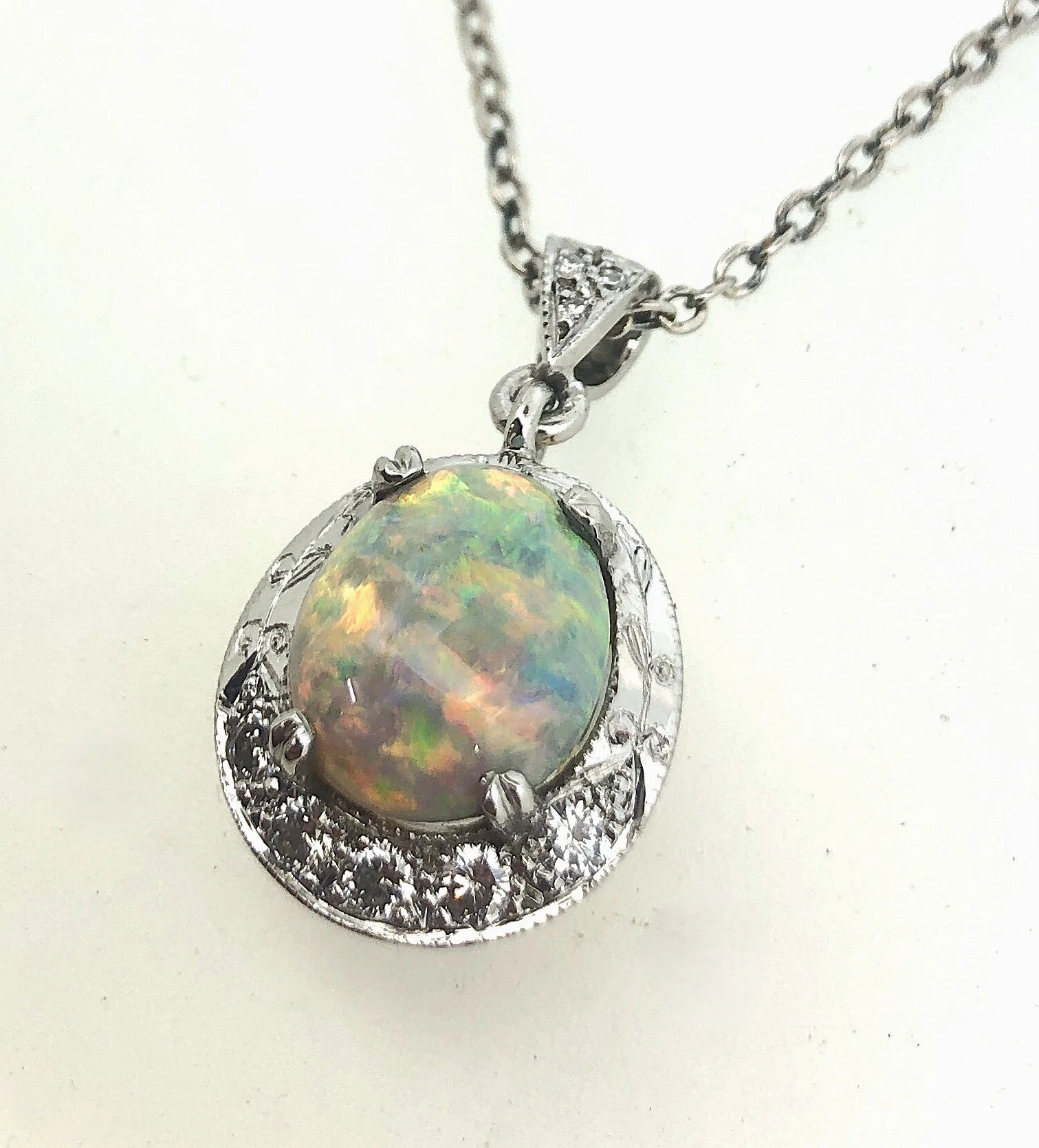 A white gold pendant necklace with a large, oval-shaped, multicolored opal gemstone surrounded by small diamonds. 