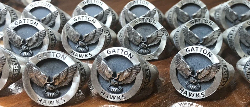 Multiple silver rings with an eagle emblem and the words "GATTON HAWKS" engraved on them, arranged on a wooden surface.