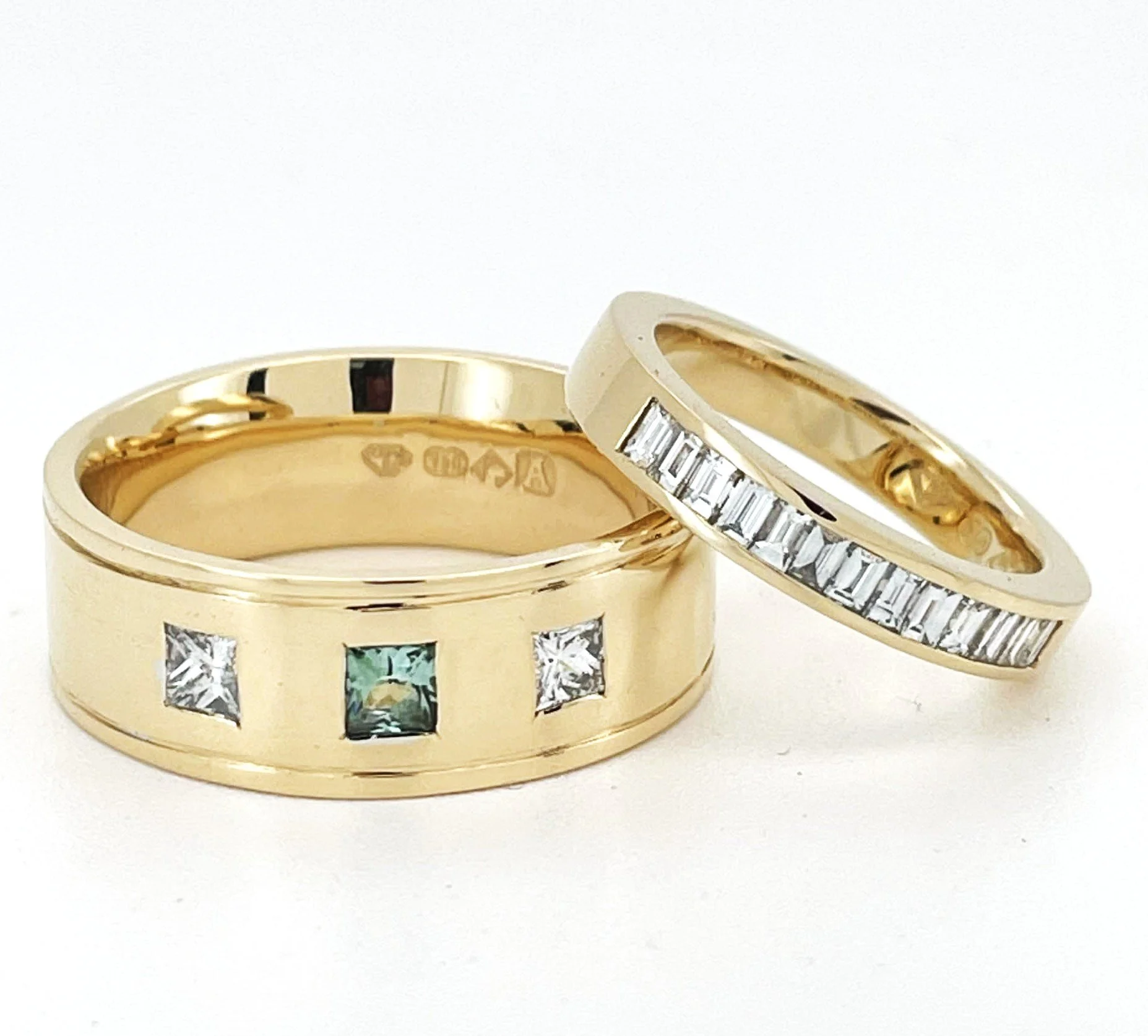 Close-up of two gold wedding rings, one with a row of small rectangular diamonds and the other with a hallmark inside. Both rings are set against a plain white background.