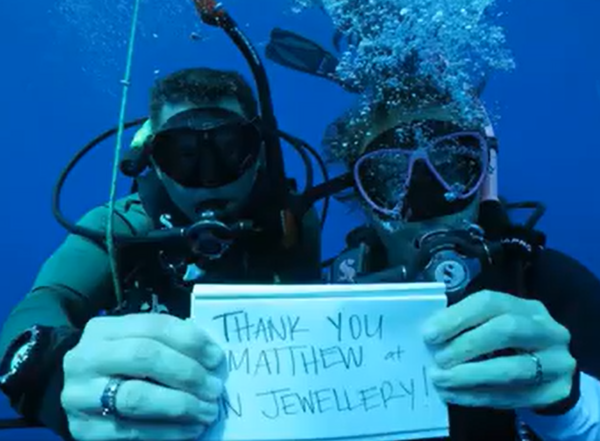 Two scuba divers underwater holding a sign that says, 'Thank you Matthew at MN JEWELLERY!'