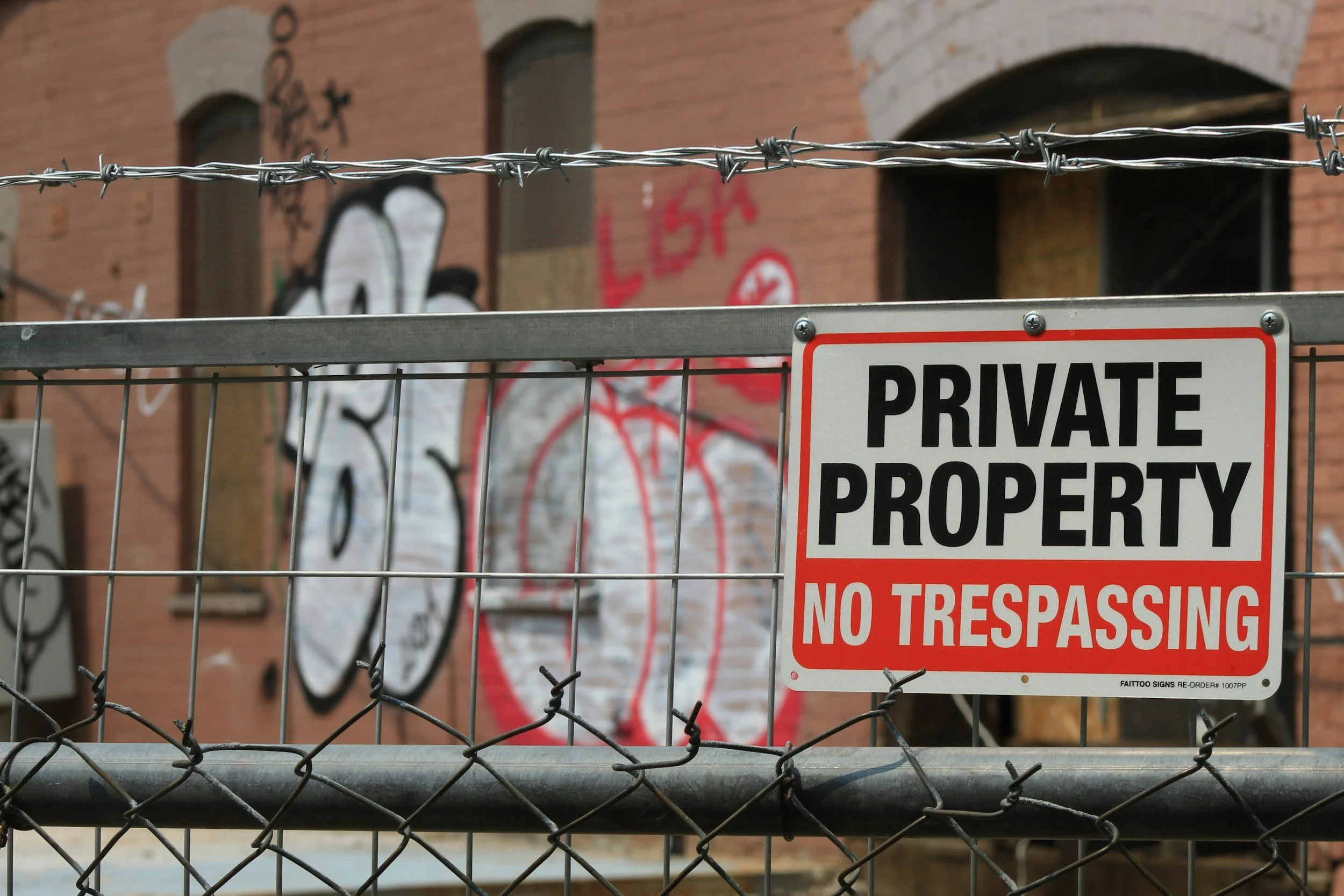 A metal fence with a 'Private Property No Trespassing' sign in front of a graffiti-covered brick building.