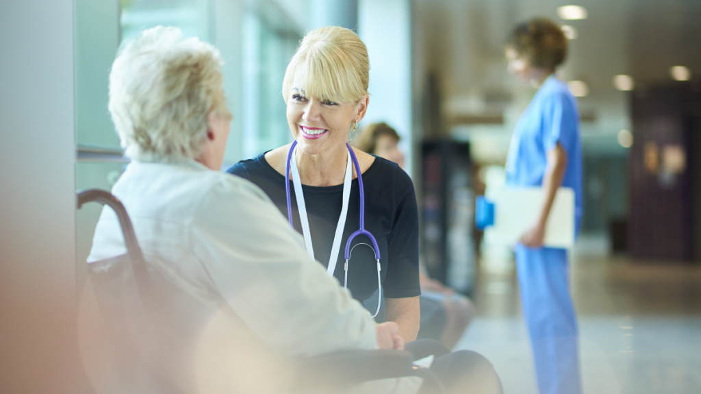 A smiling female healthcare professional talking to an elderly patient in a hospital or clinic setting, with another healthcare worker in the background.