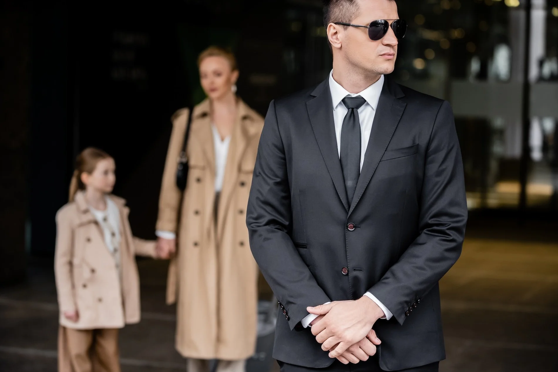 A man in a black suit and sunglasses stands in the foreground while a woman and a young girl, both wearing beige coats, hold hands in the background.