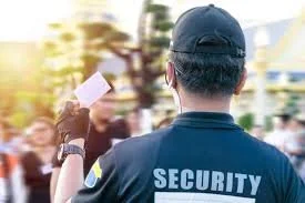 Security guard in blue uniform holding a document in an outdoor setting with people in the background.