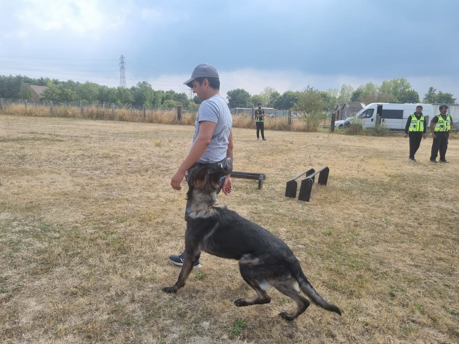 A man wearing a grey cap and t-shirt stands in a dry grassy field with a German Shepherd dog sitting next to him. In the background, there are three police officers and a white police van.
