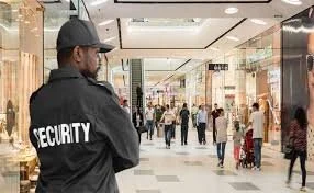 Security guard standing in a shopping mall corridor with stores and shoppers in the background.