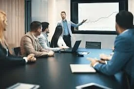 Business meeting with a man presenting financial charts on a screen to a group of four seated professionals in a conference room.