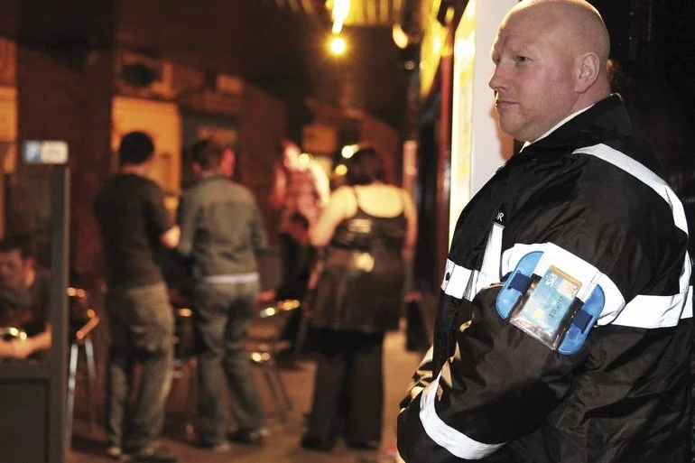 A security guard standing outside a bar or restaurant at night with people socializing in the background.