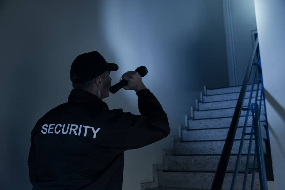 Security guard using a flashlight to look up the stairs in a dimly lit building.