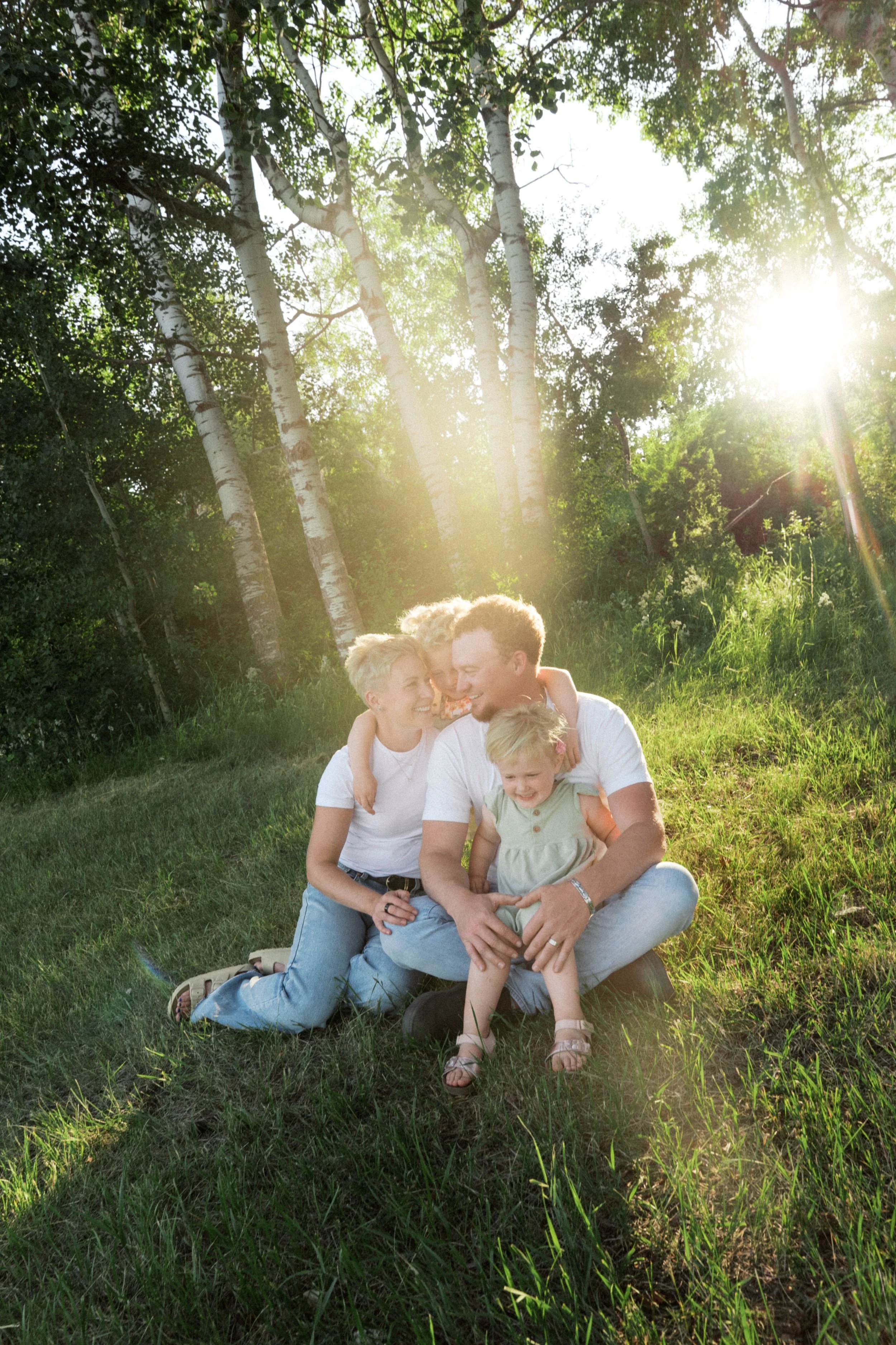 Family of four smiling and sitting on grass in a sunny park with trees in the background, enjoying a moment together.