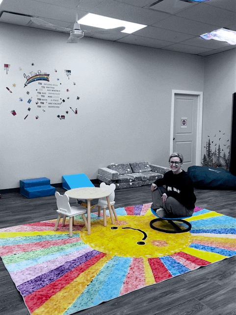 sitting on a black ring on a colorful rainbow-themed rug in a playroom. There is a small white table with a white chair, a blue lounger, a gray sofa with patterned pillows, and a large black bean bag. The room has a white wall with a decorative message and small colorful art pieces, and a white door with a winter landscape decal.