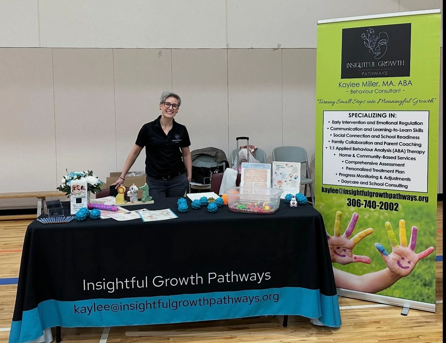 A woman stands behind a table with promotional materials and small toys, next to a green banner displaying her name as Kaylee Miller, a behavior consultant from Insightful Growth Pathways, at an informational booth in an indoor gymnasium.