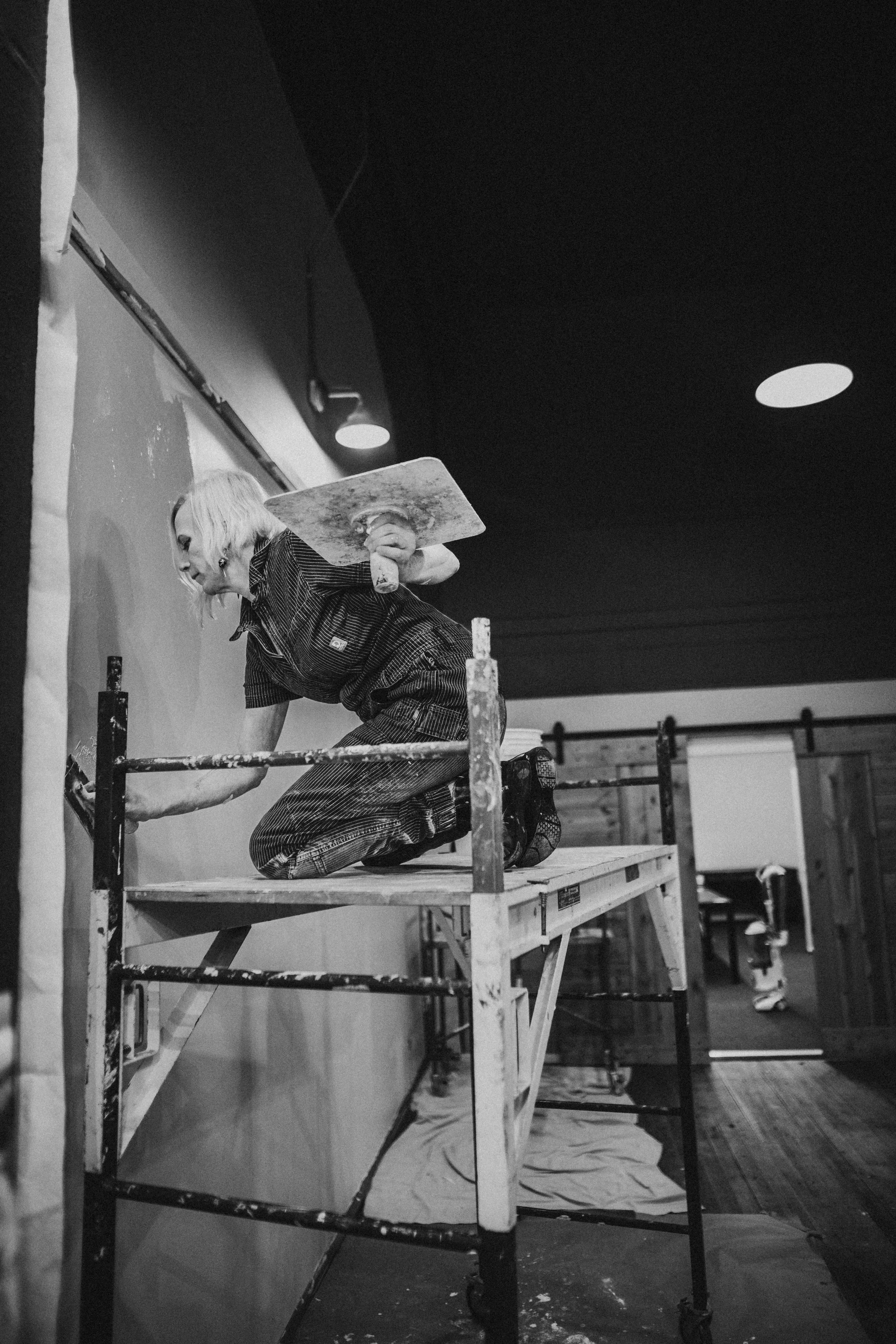 A woman working on a wall, kneeling on a scaffold, holding a trowel and a piece of plaster, with a roll of plaster in her hand, in a room with wooden walls and ceiling.