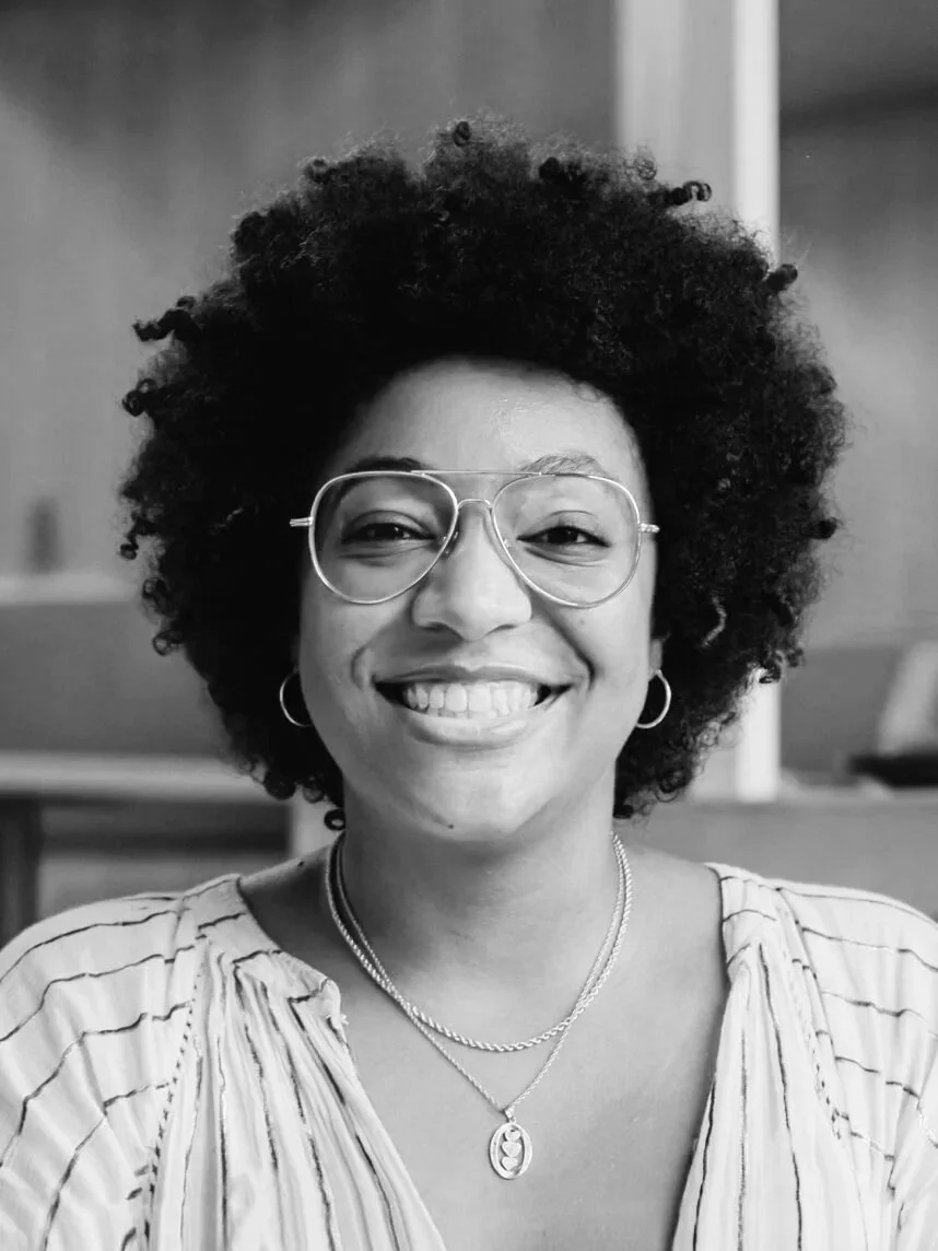 Black and white photo of a woman with curly hair, wearing glasses, earrings, and layered necklaces, smiling at the camera.