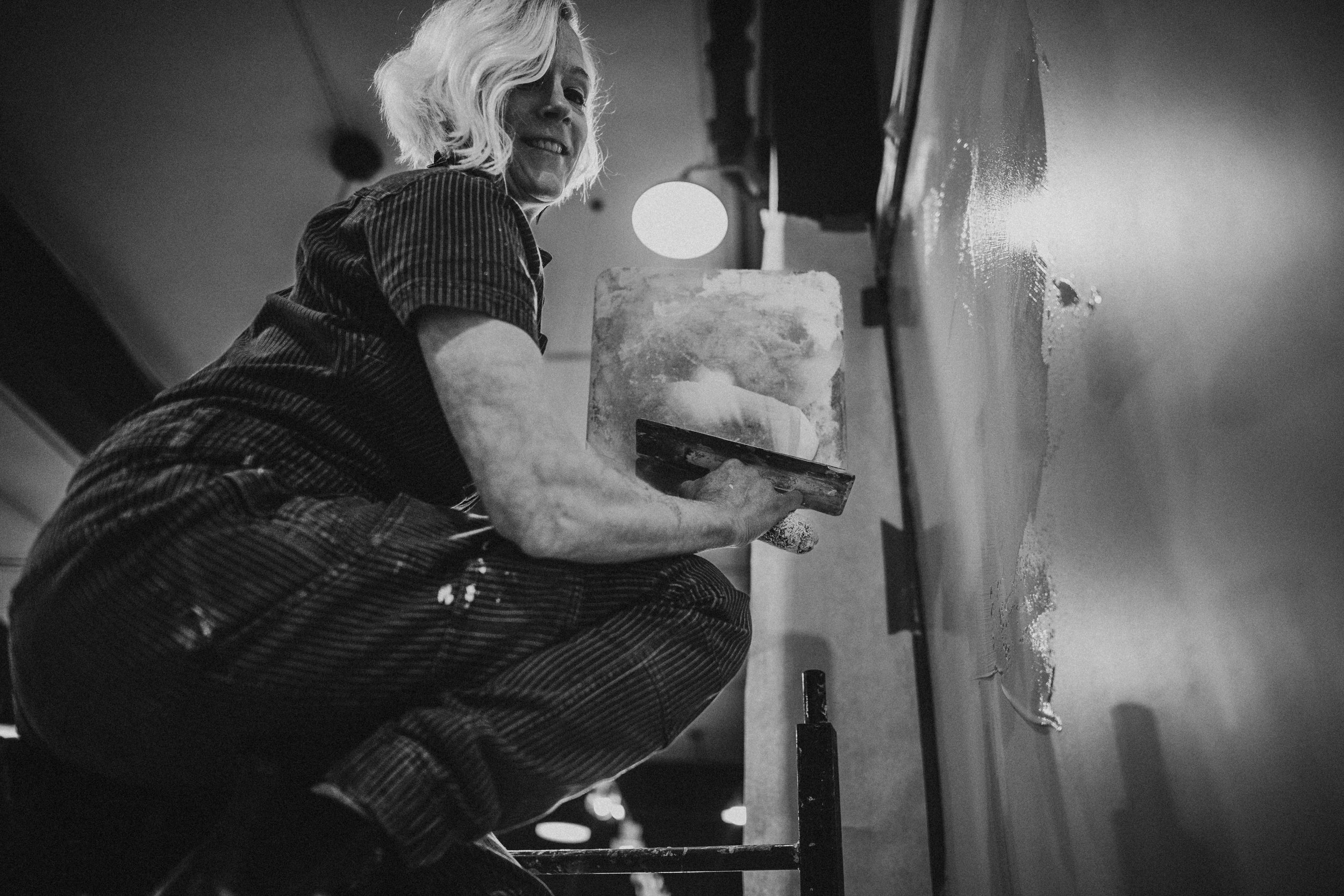 A woman with blonde hair in a short, wavy hairstyle, wearing a striped work shirt and pants, is kneeling and applying plaster to a wall with a trowel. She is smiling and looking slightly upward, with a light source behind her.