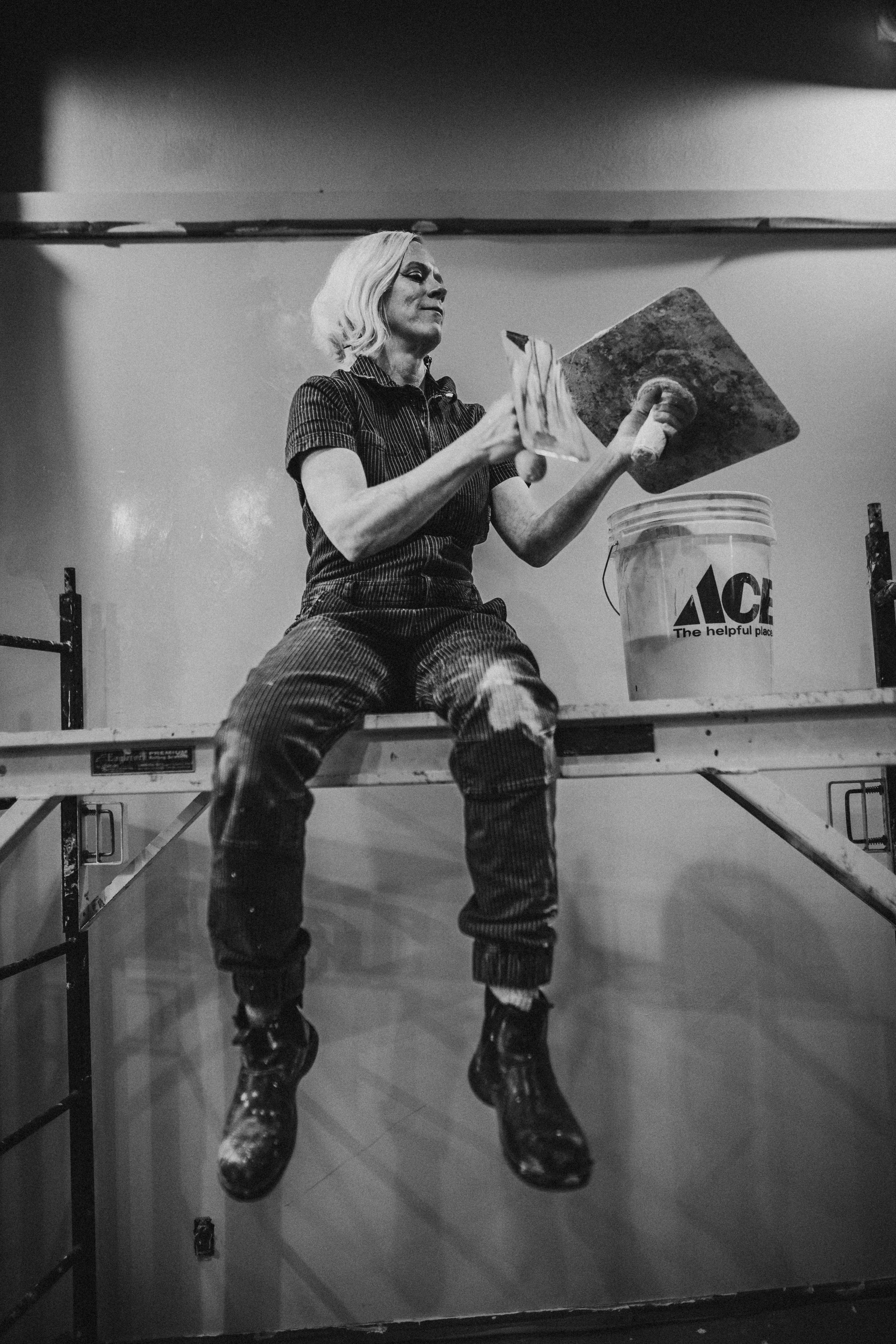 Black and white photo of a woman sitting on a workbench, holding a paintbrush and a large paint roller, with a bucket nearby, in a workshop or garage.