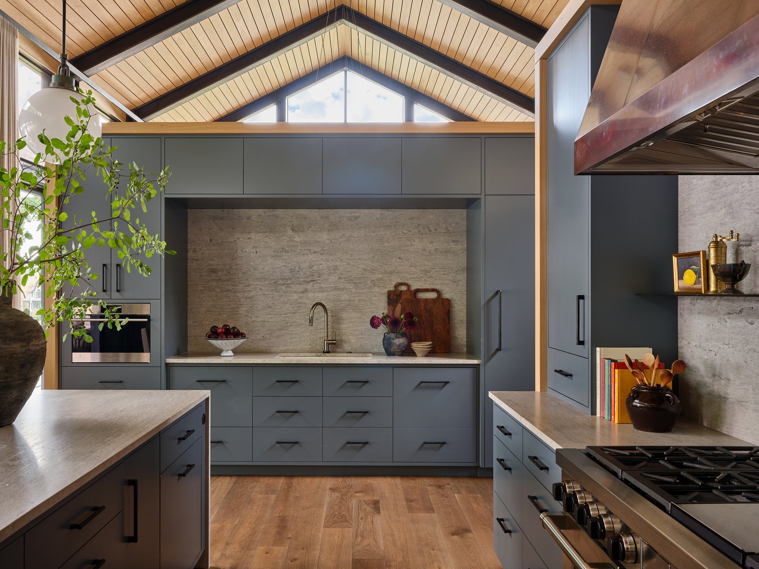 Modern kitchen with blue cabinets, a gray stone backsplash, wood accents, and stainless steel appliances, including a stove and oven. There's a potted plant on the left and decorative items on the shelves on the right.