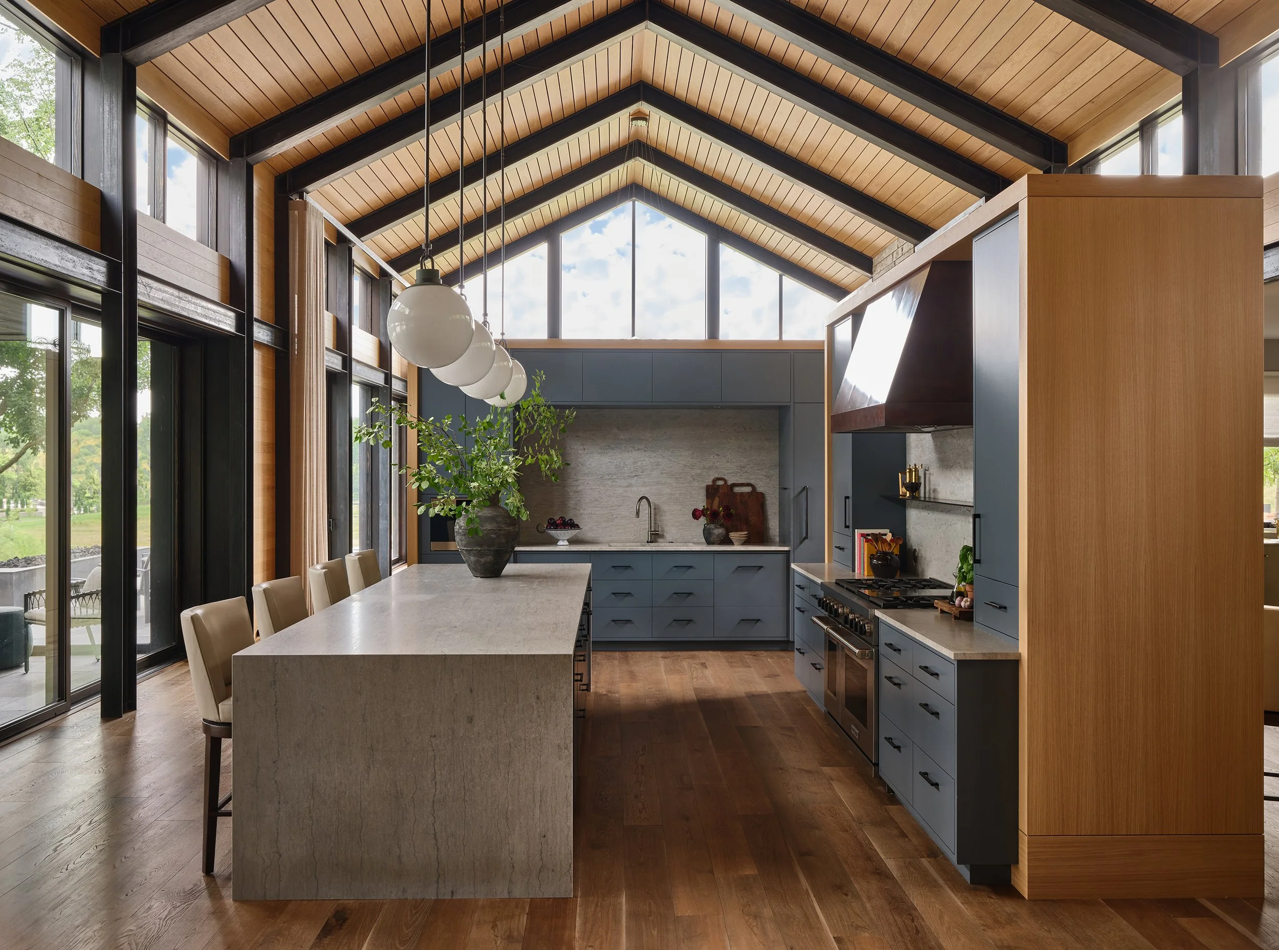Modern kitchen with high vaulted ceiling, wooden beams, large windows, blue cabinets, island with beige countertop, four chairs, and pendant lights, with a view of outdoors.