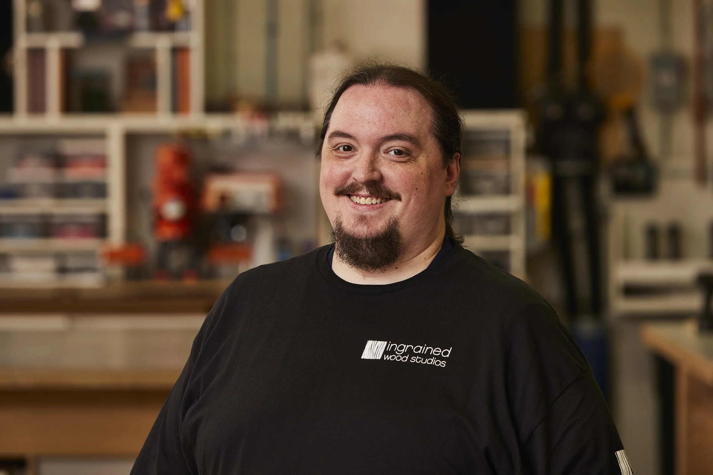 A smiling man with dark hair, a goatee, and mustache wearing a black shirt with 'inspired wood studios' logo in a woodworking workshop.