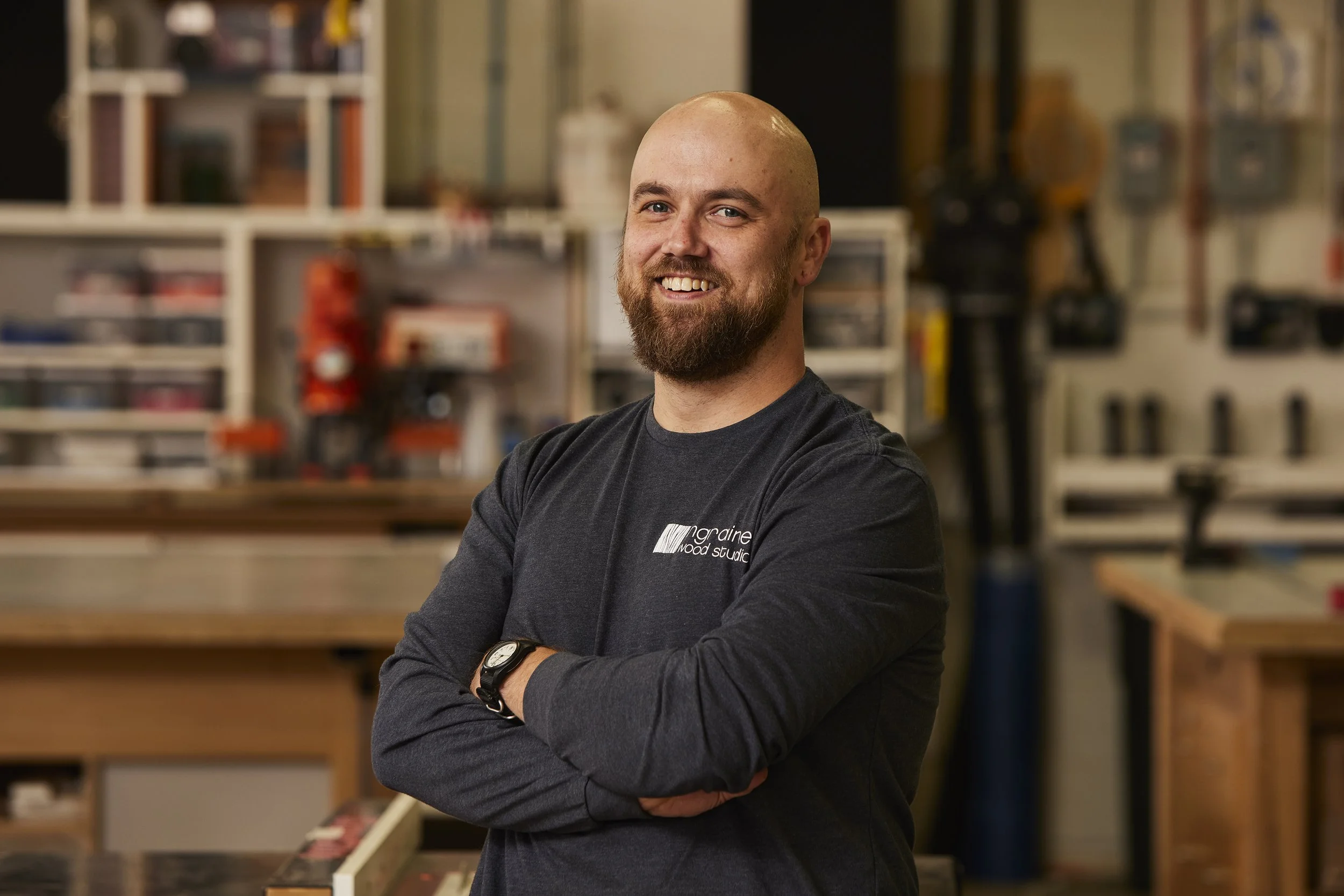 A smiling man with a beard and bald head stands with his arms crossed in a woodworking or craft workshop. Background features shelves with various tools, supplies, and materials.