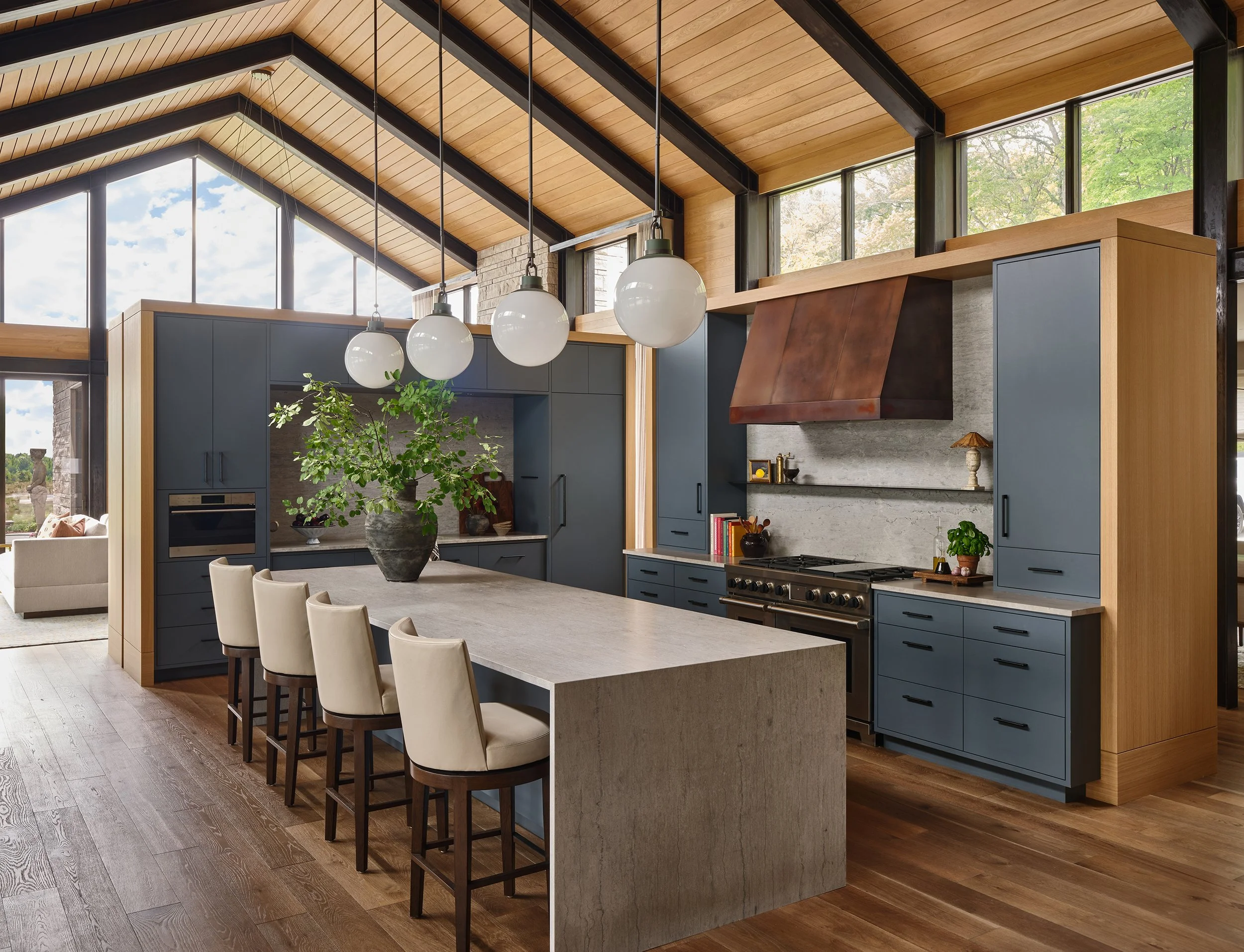 Modern kitchen with blue cabinets, a wooden ceiling, a large island with white chairs, a large plant in a pot, and a stove with a rust-colored range hood.