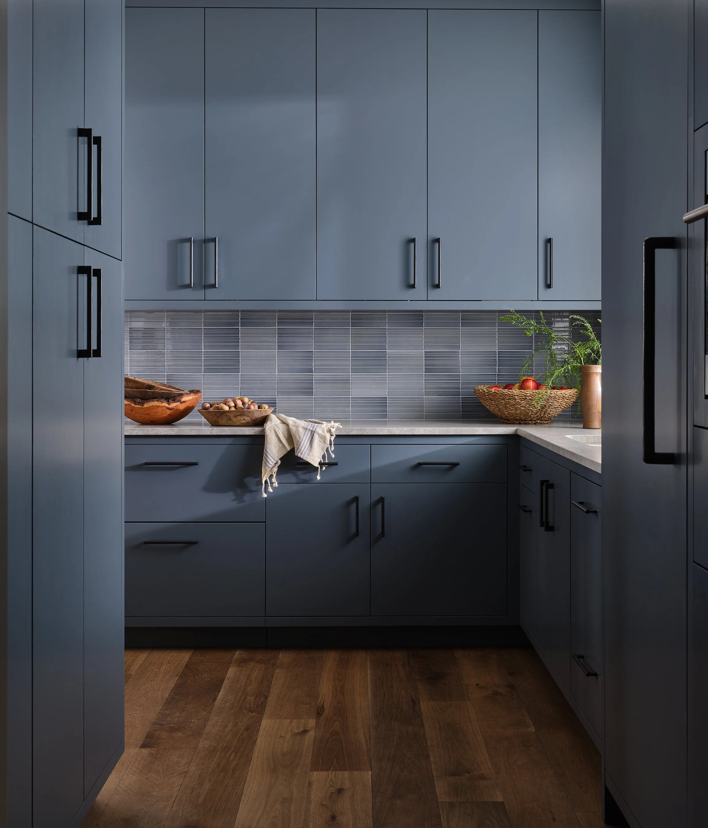 Modern kitchen with blue cabinets, wood flooring, and a gray tile backsplash, featuring bowls of nuts, a basket of apples, and a potted plant.
