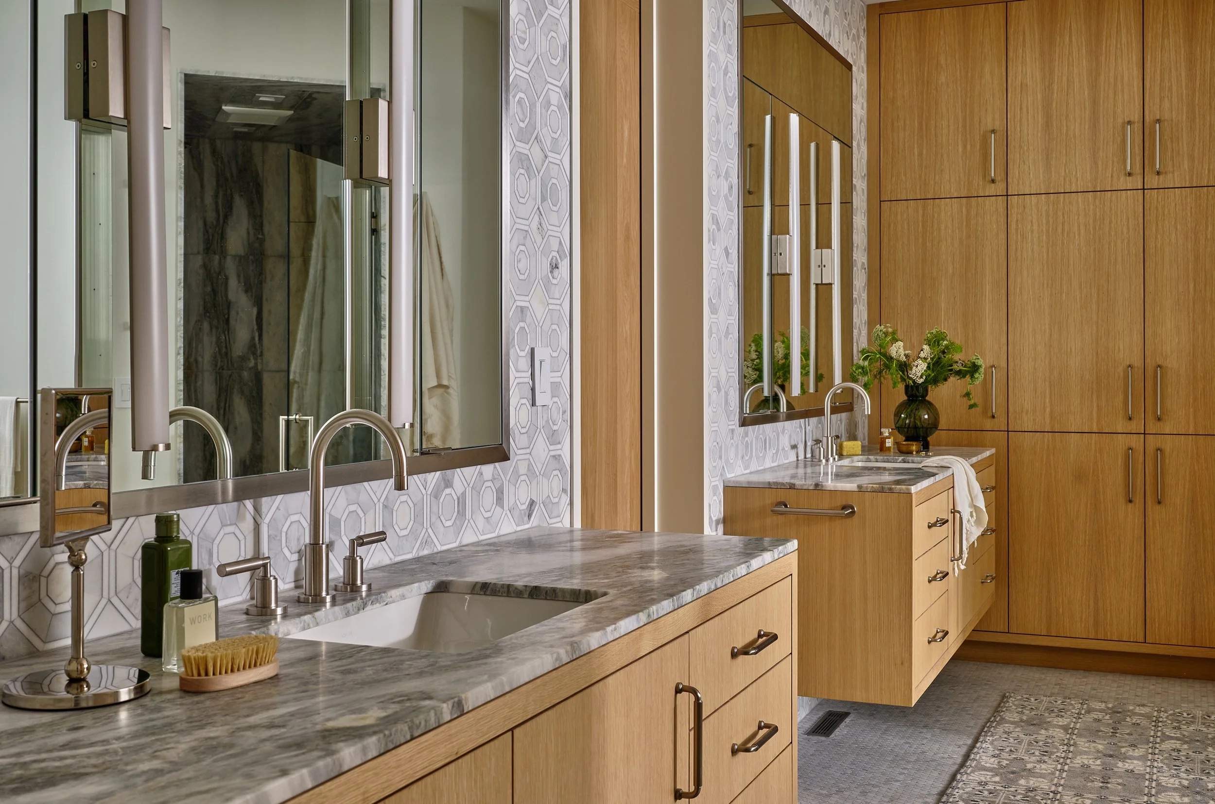 Bathroom with two marble countertops, wooden cabinets, oval mirrors, sinks, and a towel, decorated with a vase of flowers and patterned wallpaper.