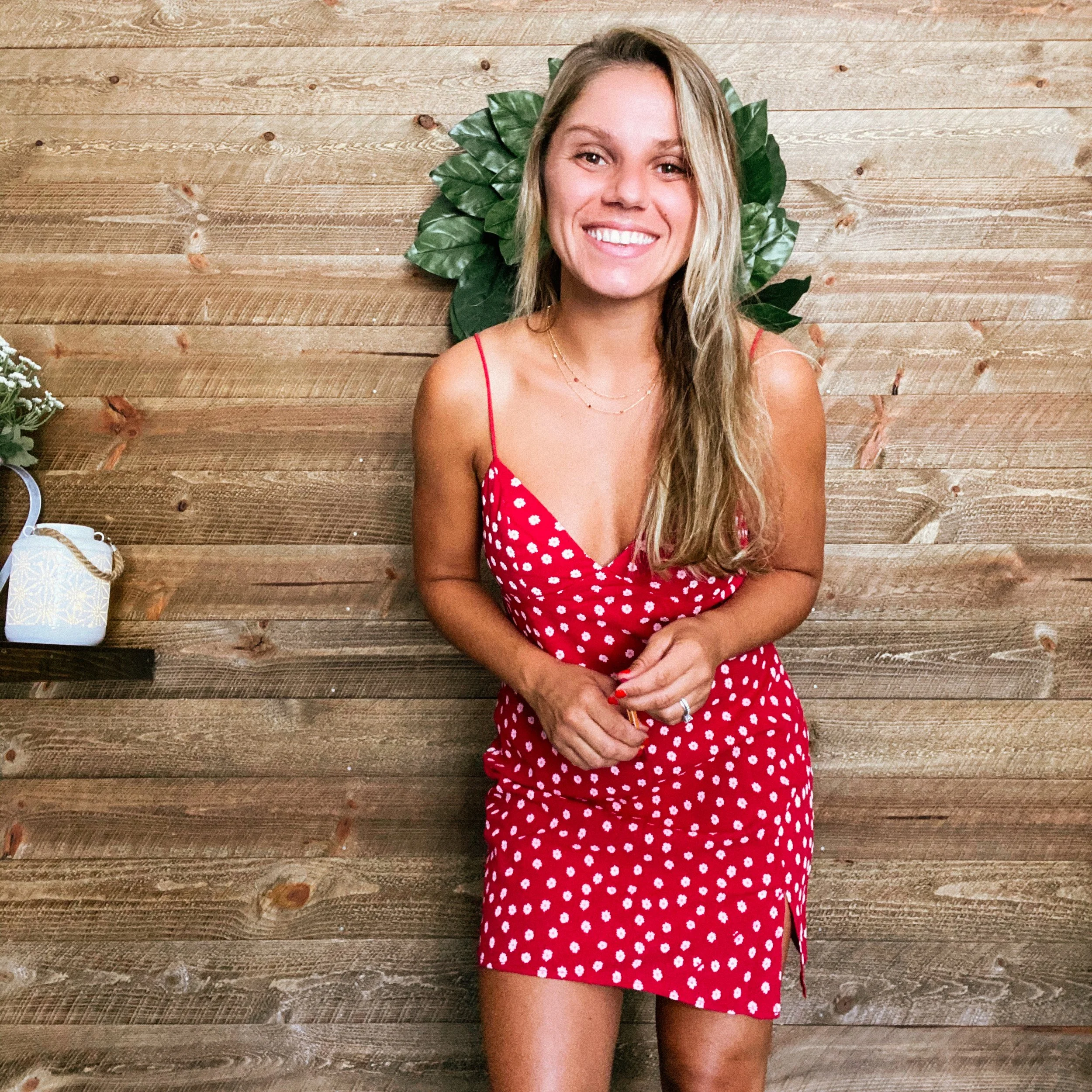 A smiling woman with long blonde hair, wearing a red polka dot dress with spaghetti straps, poses in front of a wooden wall with green leaves behind her.