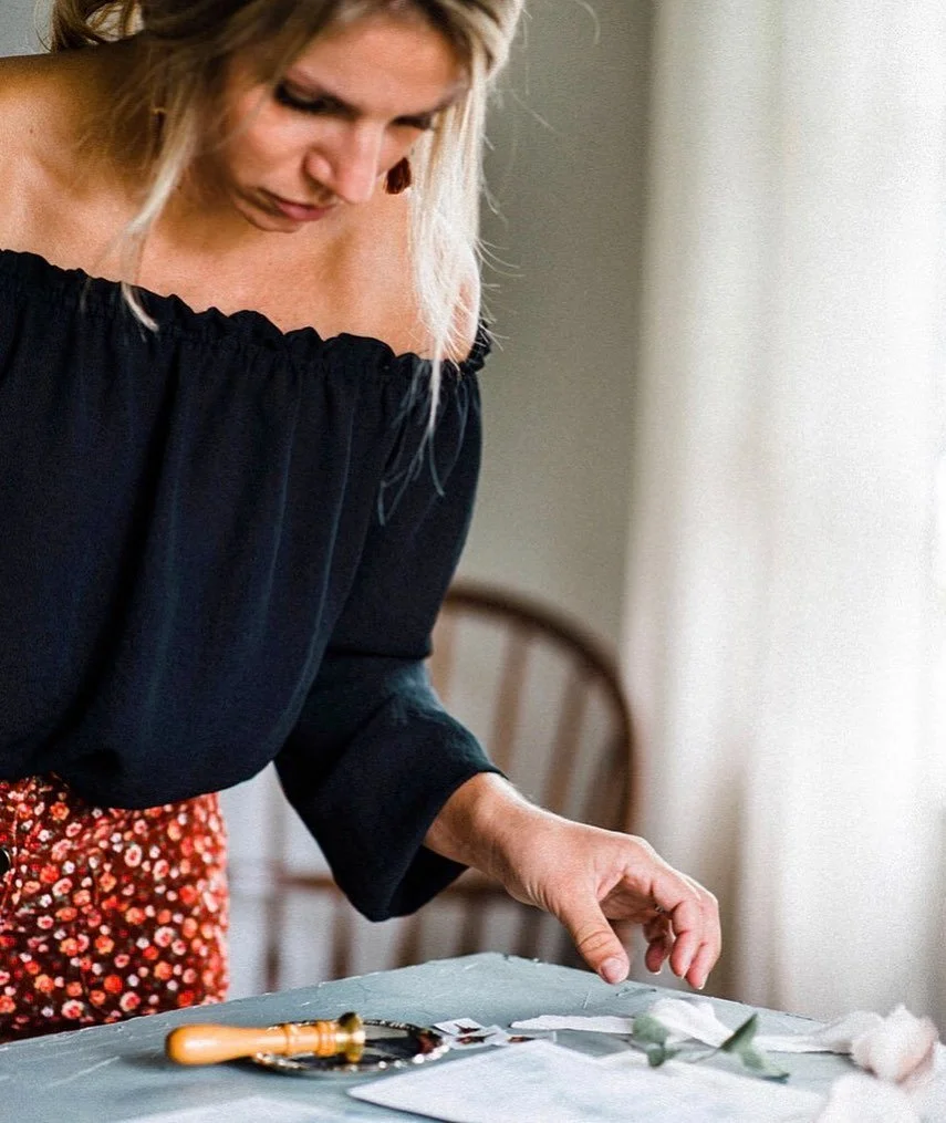 Sophie. Sincerely Sophie assembling a flat lay of a custom wedding suite. A woman wearing a black off-the-shoulder top and a floral skirt is arranging small flower arrangements and wedding stationery on a table.