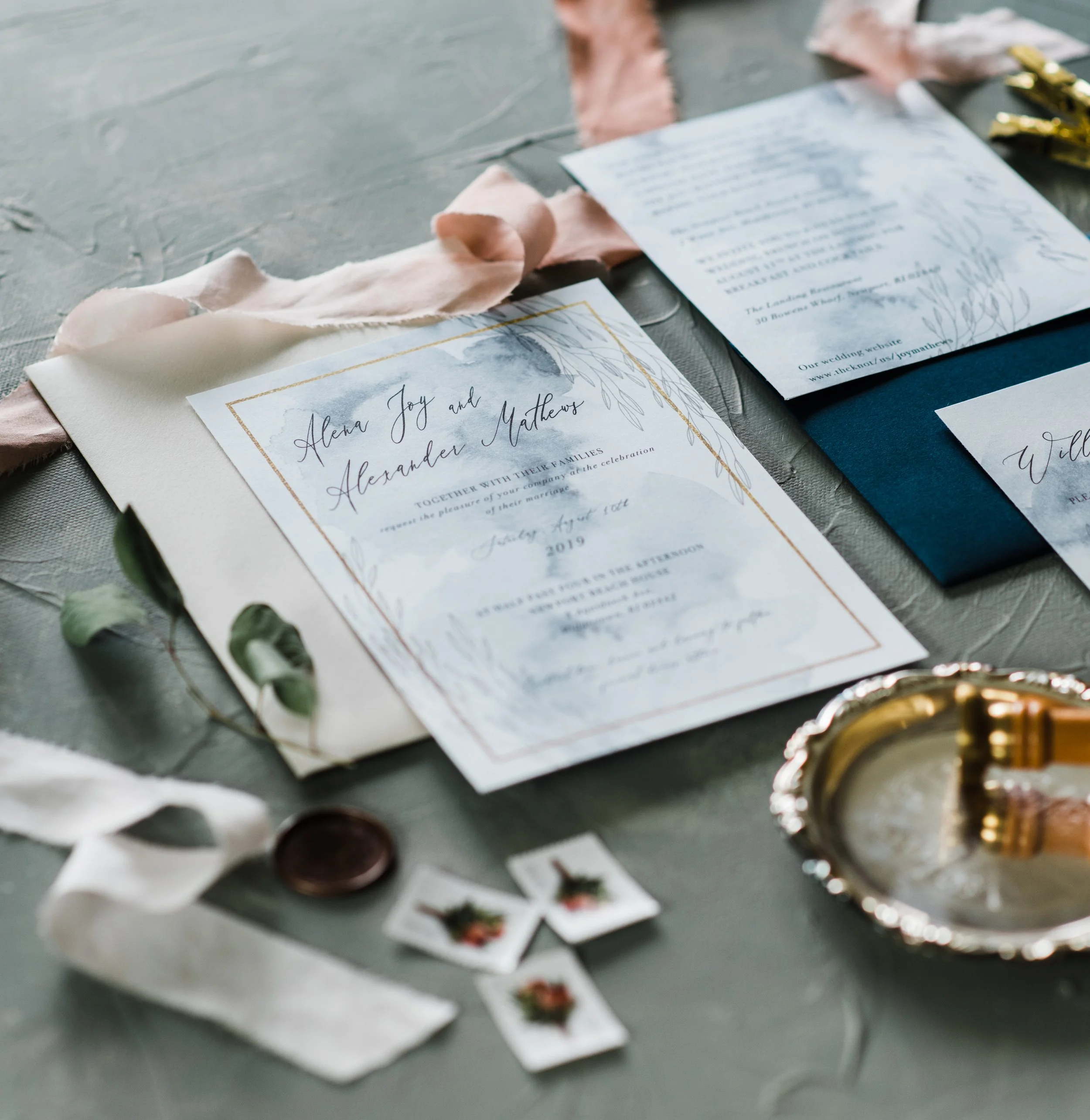 Wedding invitation with floral design, along with a blue card, small matching gift tags, a gold ring, and decorative ribbons on a textured surface.