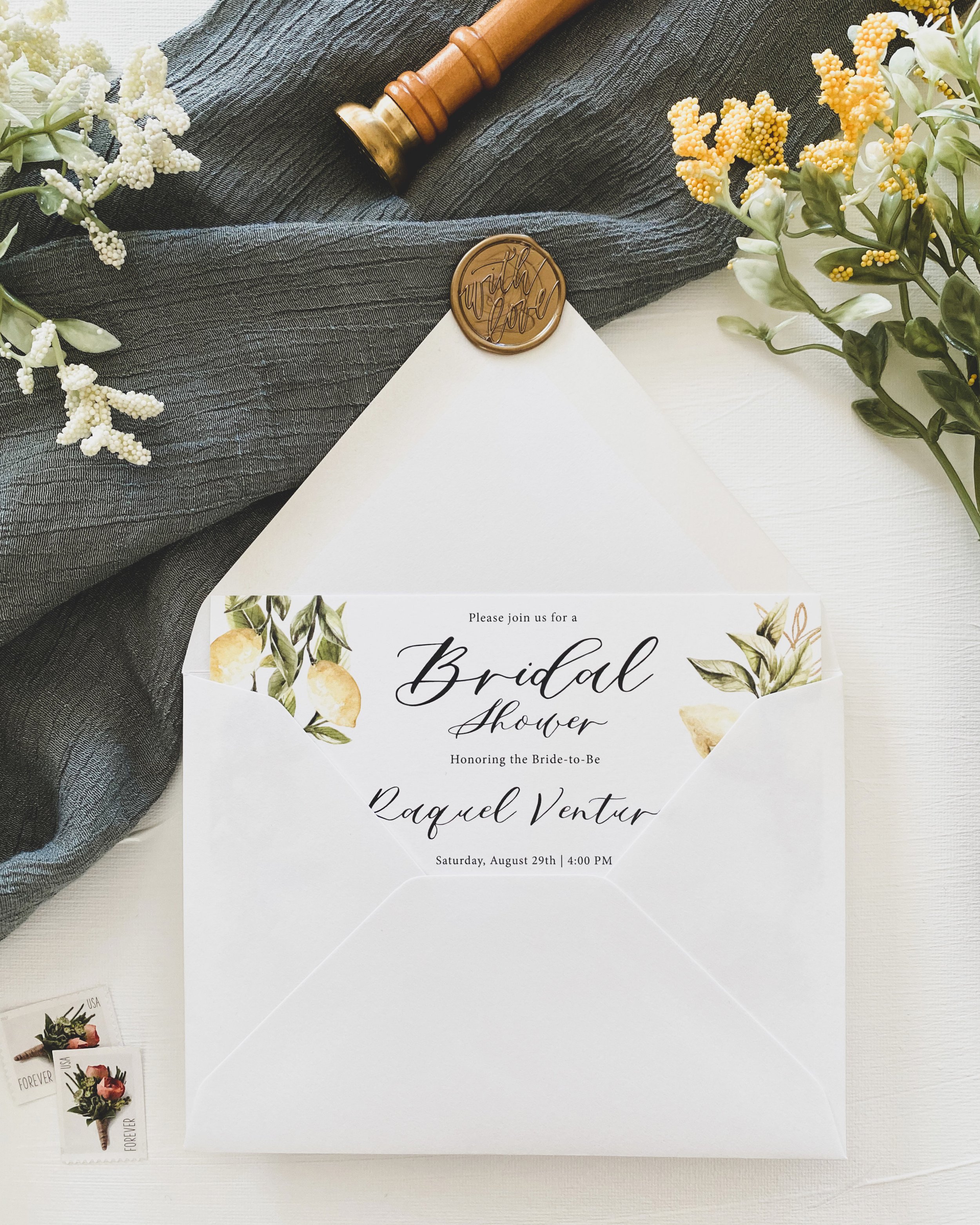 A bridal shower invitation inside a white envelope with a decorative seal, surrounded by flowers and lemons for a summer event in Rhode Island. Forever US postage. 