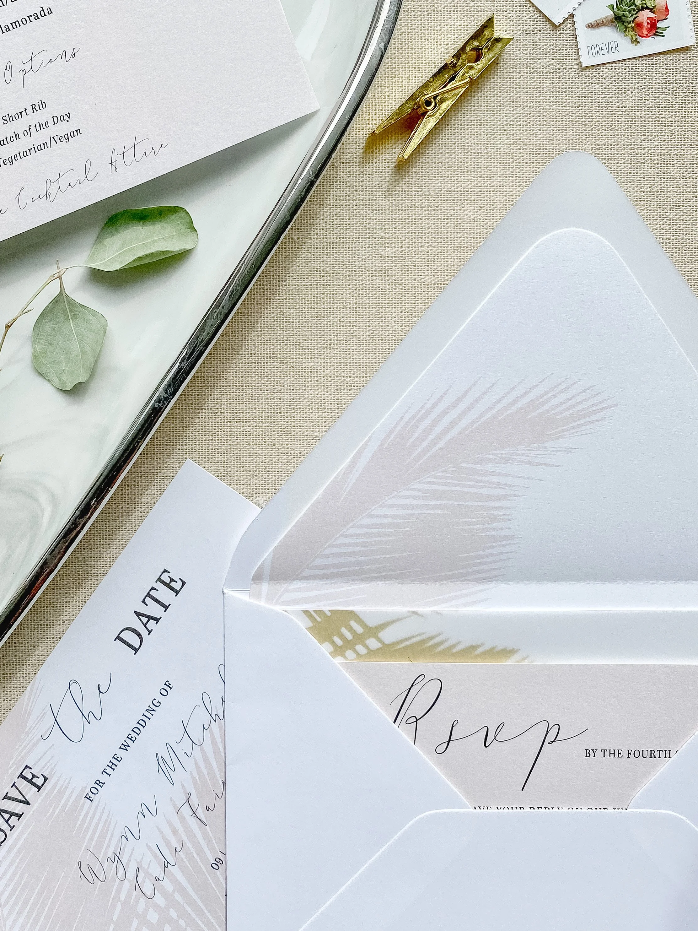 Wedding invitations with a palm leaf design, gold clip, leaf decoration, and a tray with papers and a flower.