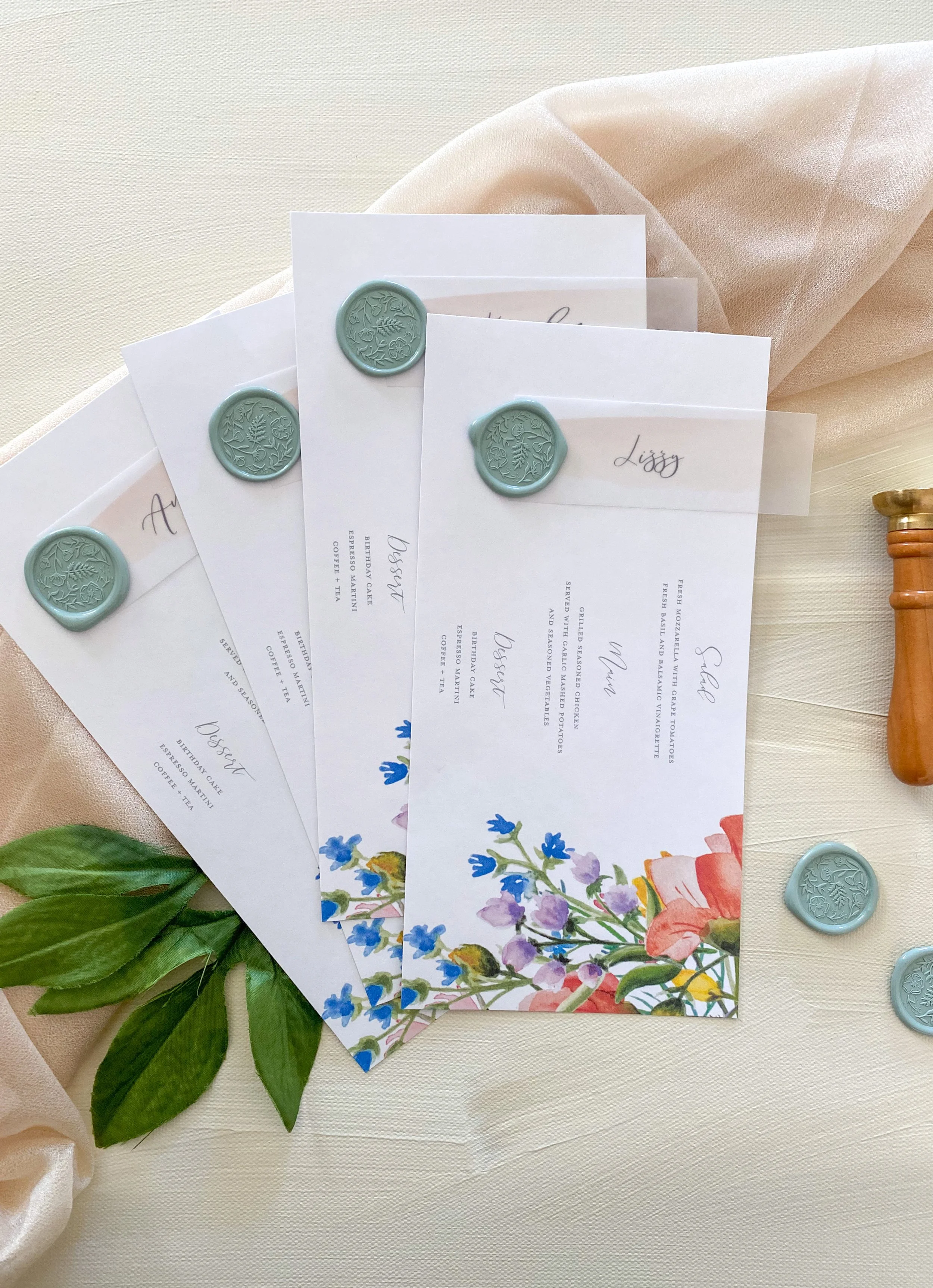 Three wedding menus with blue wax seals, decorated with floral designs at the bottom, placed on a cream-colored tablecloth with a soft pink fabric, green leaves, and some scattered blue and gold wax seals.