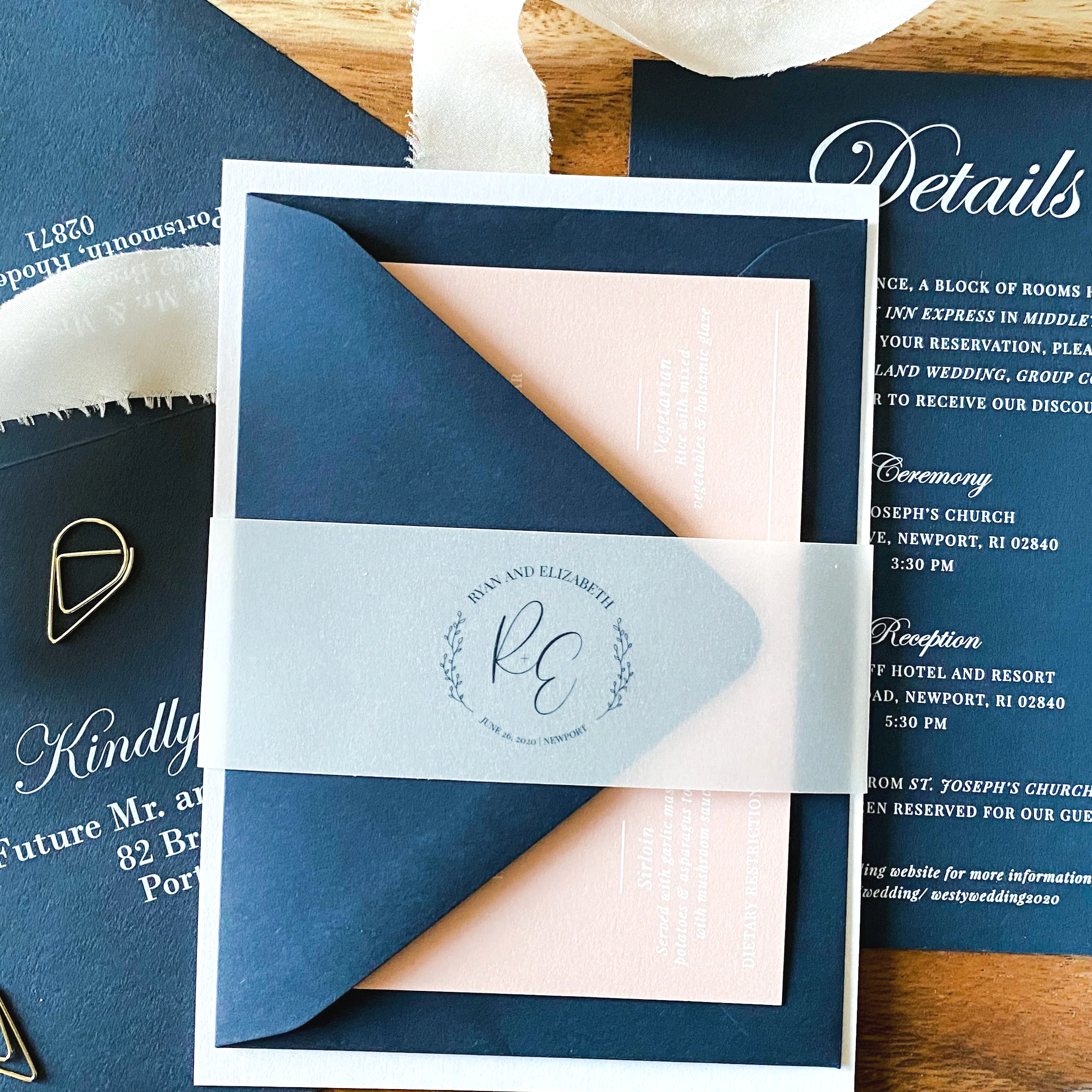 Wedding invitation suite including an outer navy blue envelope, a pale pink insert, and a white band with a monogram of 'R E' and the names Ryan and Elizabeth. The suite is decorated with a gold paperclip, a piece of cream-colored ribbon, and other wedding stationery in the background.