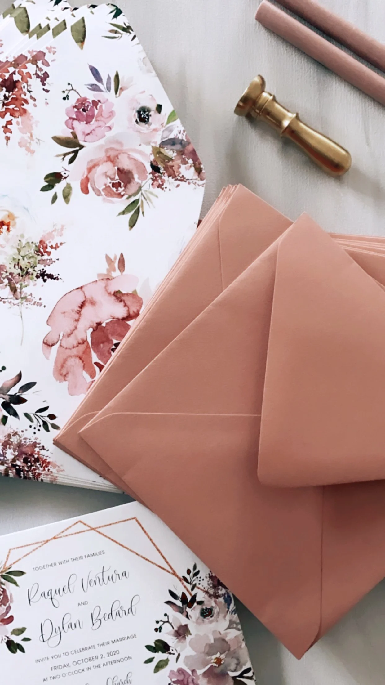 A floral wedding invitation with pink envelopes, and floral envelope liners, printed on white paper with pink, purple, and green watercolor flowers; a gold wax seal stamp; pink napkins.