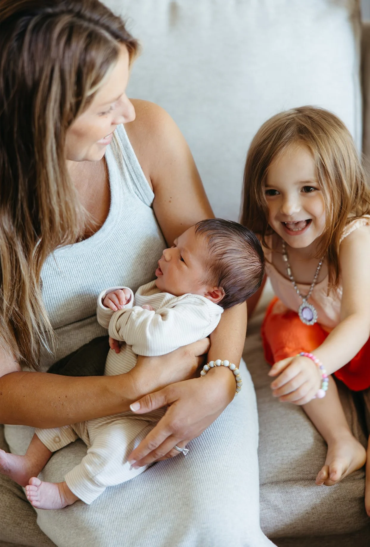 A woman holds a newborn baby while a little girl smiles and leans in, showing affection and curiosity.