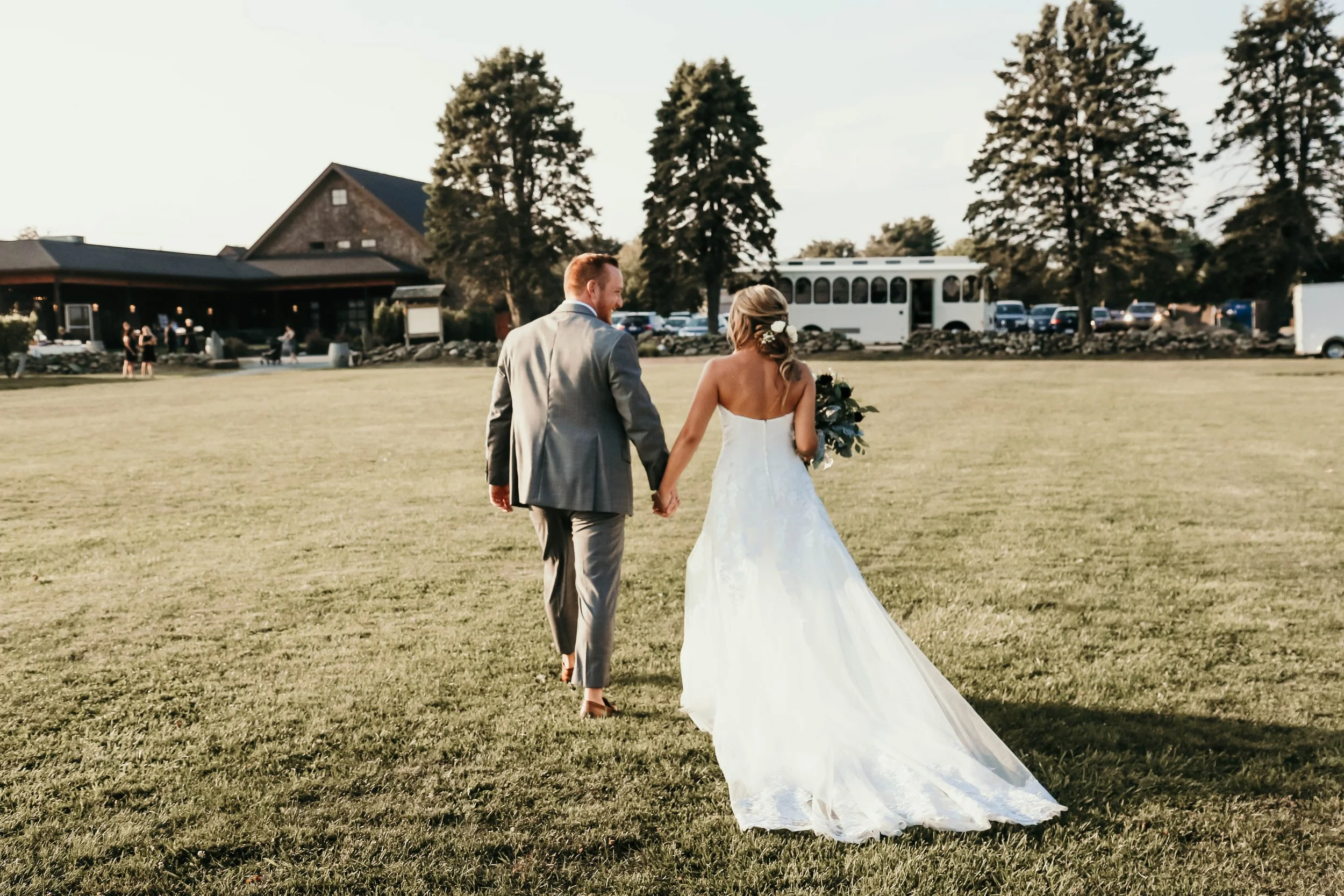 A bride and groom walking hand-in-hand on a grassy field during their wedding at sunset, with guests and trees in the background.